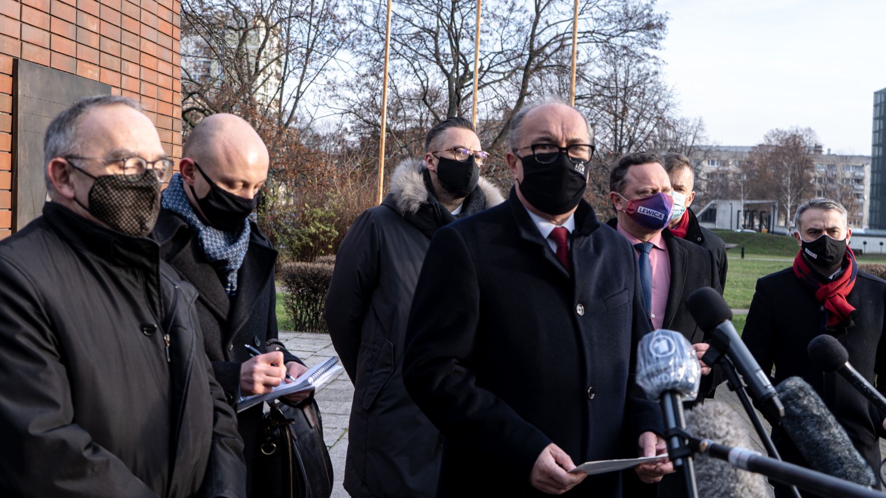 Grupa mężczyzn w maskach i zimowych płaszczach stoi na zewnątrz podczas konferencji prasowej, a jeden z nich w czarnym płaszczu i czerwonym krawacie przemawia do mikrofonów.