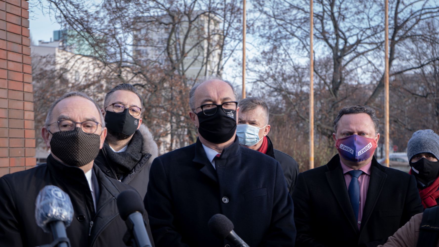 Grupa osób w maseczkach stoi na zewnątrz przed mikrofonami podczas konferencji prasowej.