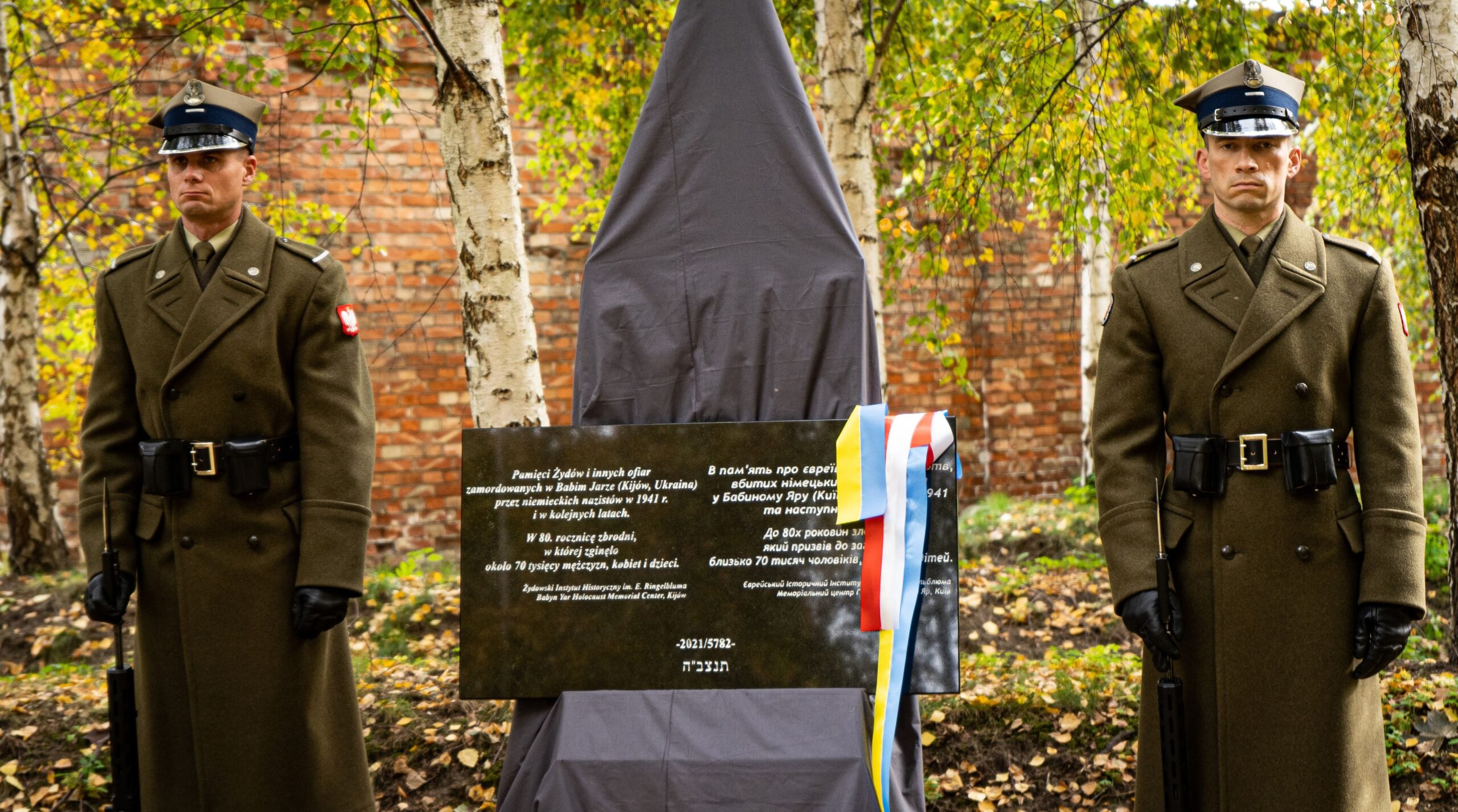 Dwóch żołnierzy w wojskowych mundurach stoi po obu stronach zasłoniętej tkaniną tablicy upamiętniającej ofiary masowych egzekucji w Babim Jarze podczas uroczystości.