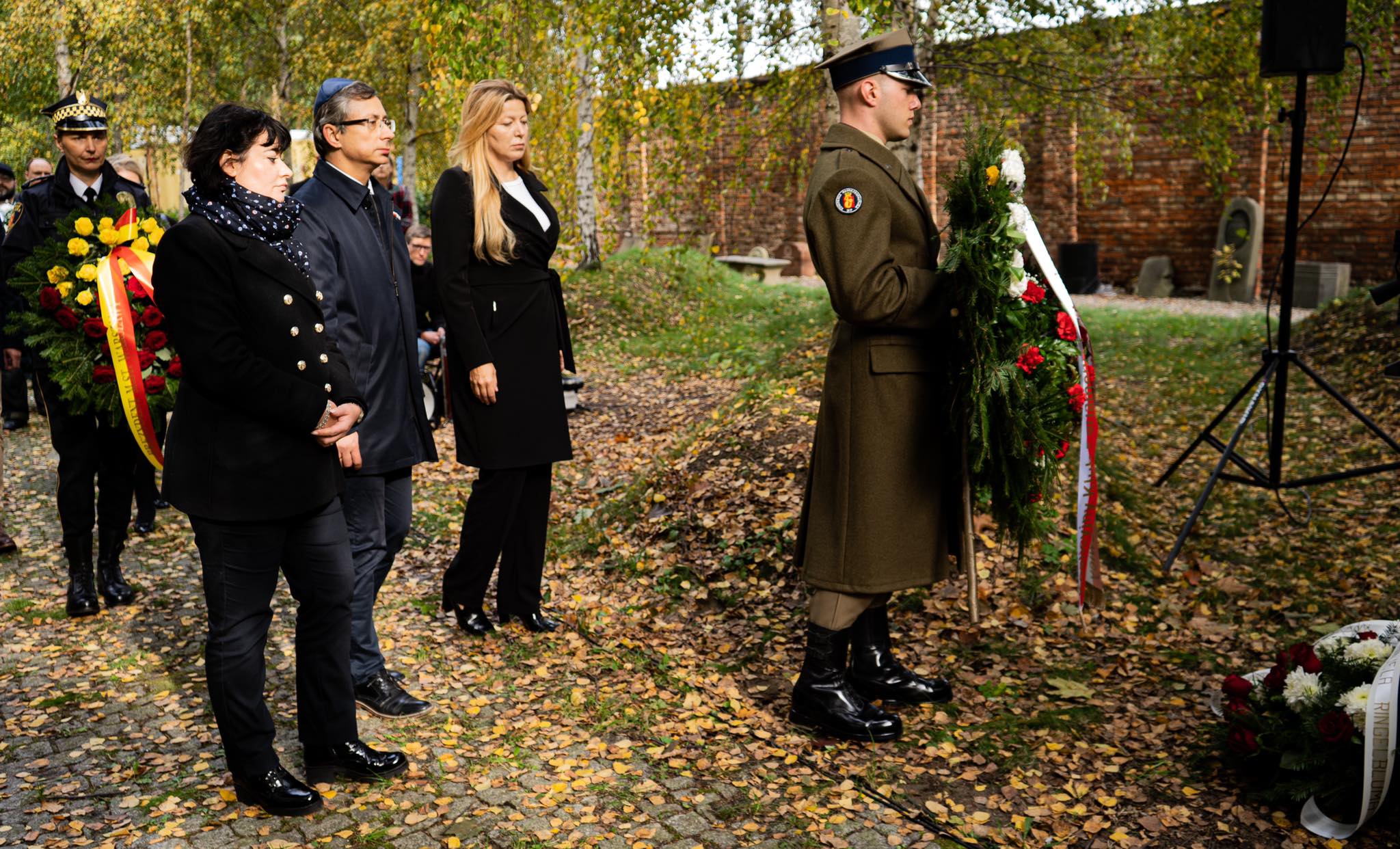 Uroczystość upamiętniająca ofiary masowych egzekucji w Babim Jarze: grupa osób w eleganckich ubraniach stoi w milczeniu, a żołnierz w mundurze trzyma wieniec podczas ceremonii w jesiennym parku.
