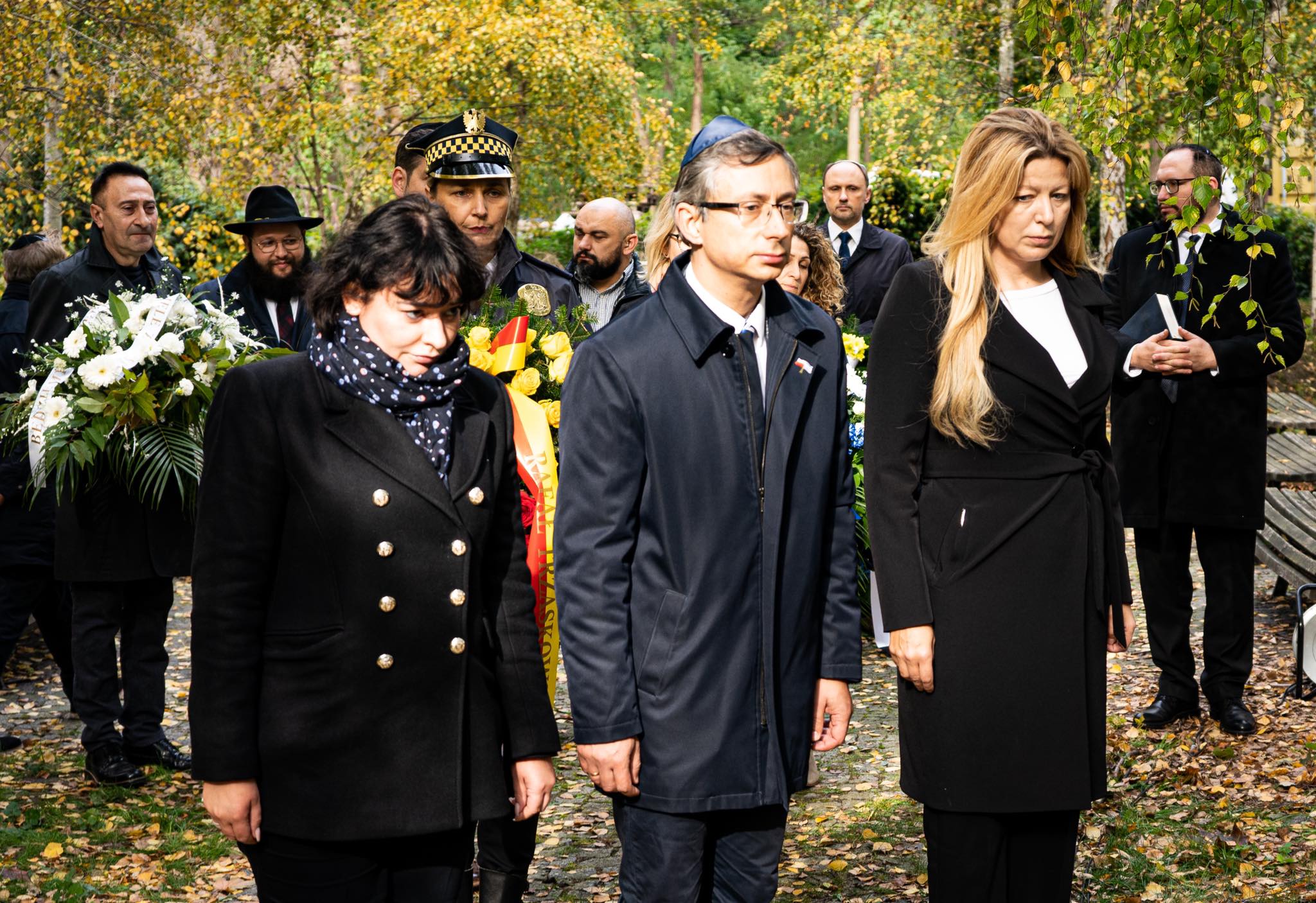 Na zdjęciu widoczna jest grupa ludzi w eleganckim stroju, z poważnymi wyrazami twarzy, uczestniczących w ceremonii upamiętniającej w otoczeniu drzew i kwiatów.