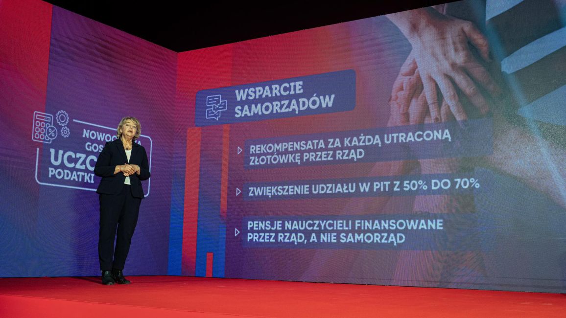 Kobieta w czarnym garniturze stoi na czerwonej scenie podczas konwencji Nowej Lewicy, a na ekranie w tle widoczne są slajdy z postulatami gospodarczymi.