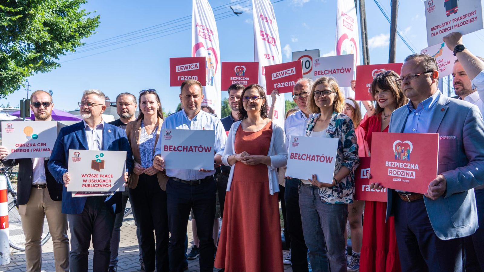Grupa ludzi na demonstracji politycznej trzyma plakaty z napisem Bełchatów i hasłami Lewica razem, na tle flag i banerów.