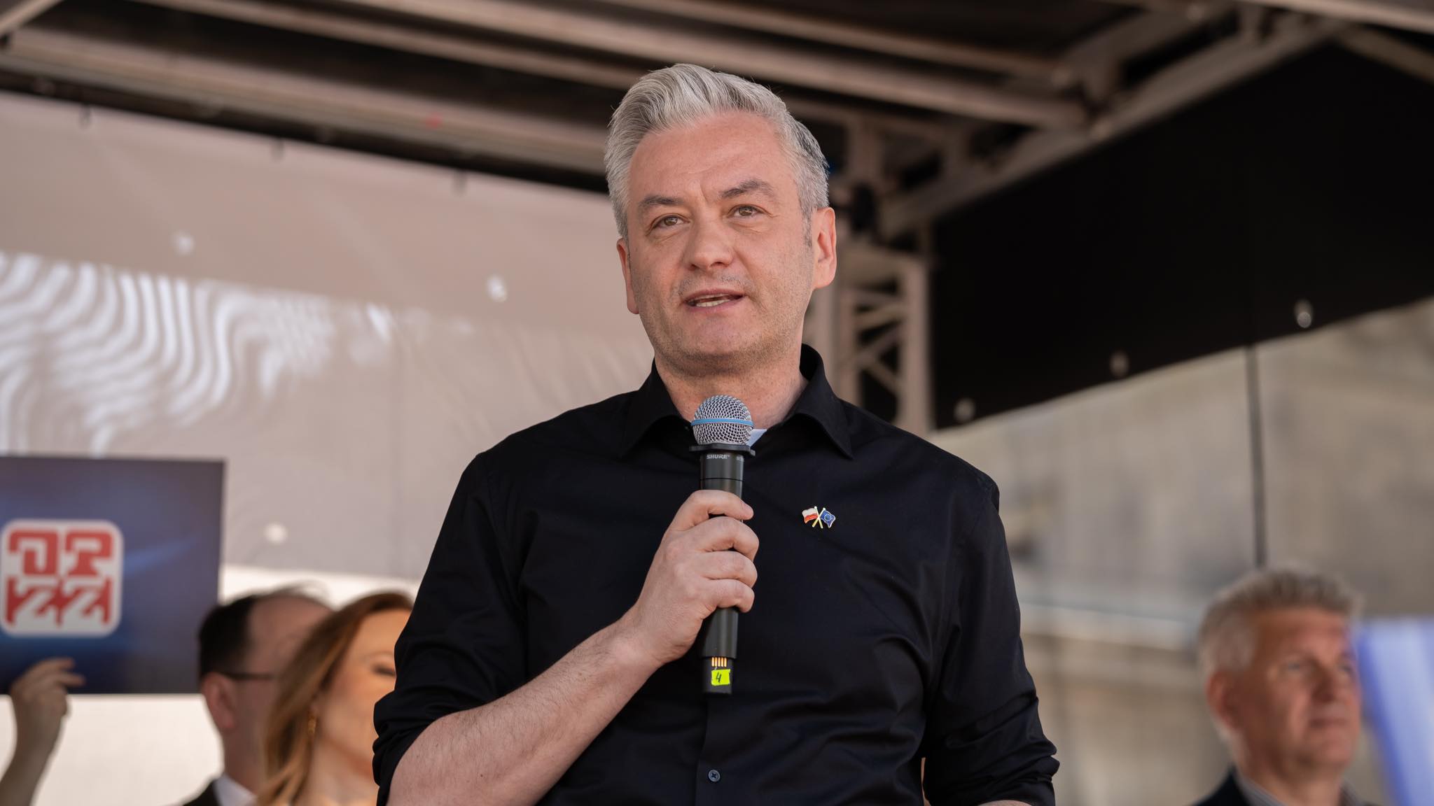 Mężczyzna z siwymi włosami w czarnej koszuli przemawia do mikrofonu na scenie podczas Święta Pracy 2024.