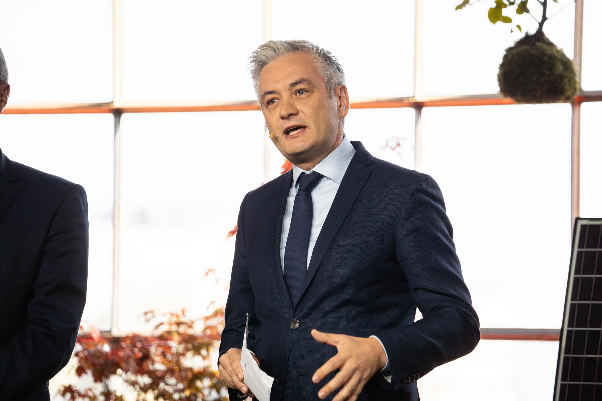 Mężczyzna w garniturze przemawia podczas wydarzenia, stojąc przed oknem z roślinami i panelami słonecznymi.