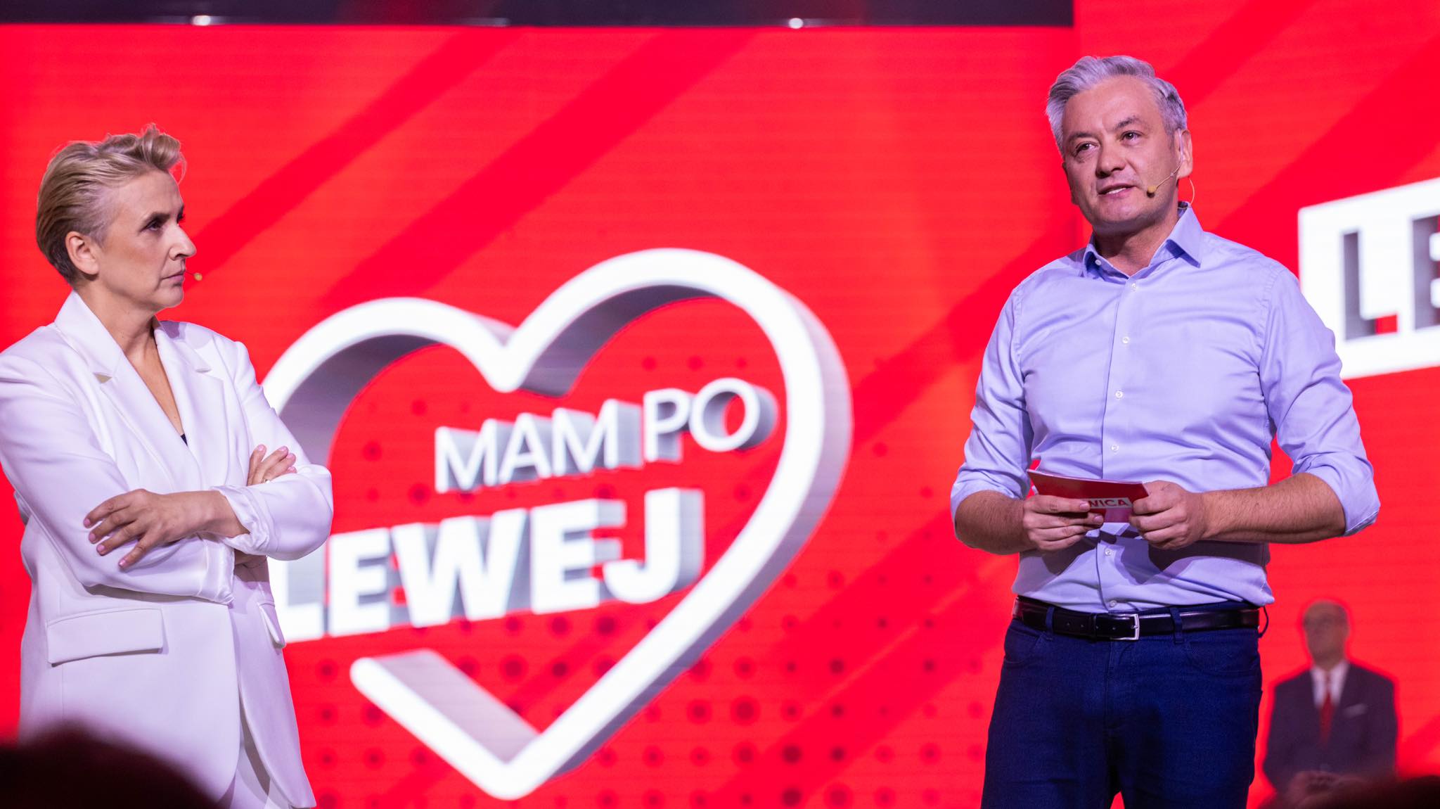 Dwóch prowadzących stoi na scenie na czerwonym tle z dużym sercem i napisem „MAM PO LEWEJ”.