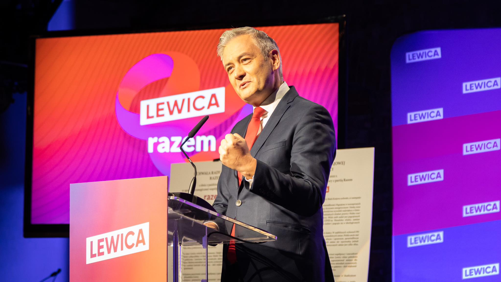 Na zdjęciu widać mężczyznę w garniturze, który z pasją przemawia przy mównicy, z logo partii Lewica w tle.