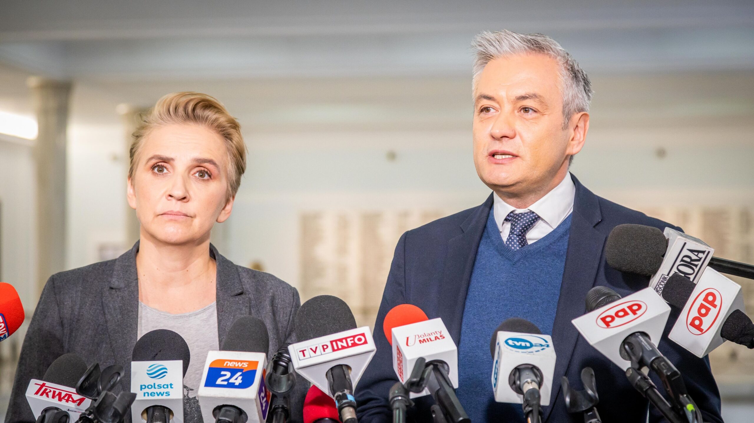 Na zdjęciu widać dwoje polityków stojących przed mikrofonami podczas konferencji prasowej.