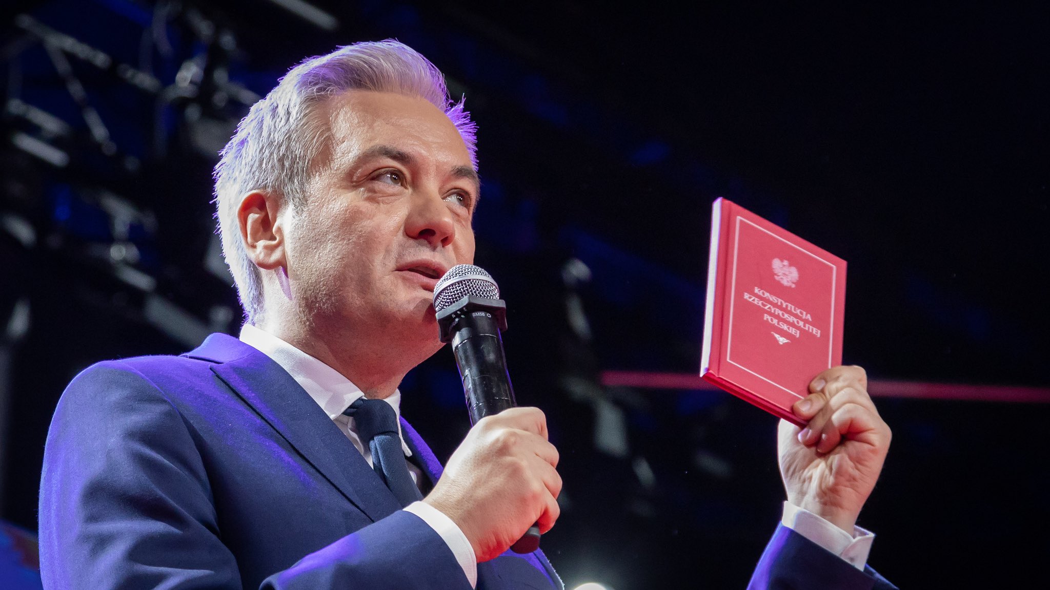 Mężczyzna w garniturze trzyma mikrofon w jednej ręce i otwartą konstytucję w drugiej, przemawiając na tle sceny.