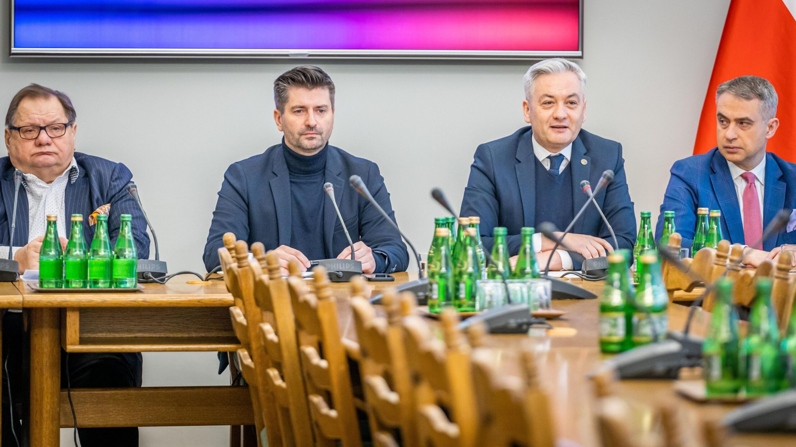 Czterech mężczyzn w garniturach siedzi przy stole, na którym znajdują się butelki zielonej wody, podczas spotkania w sali konferencyjnej.