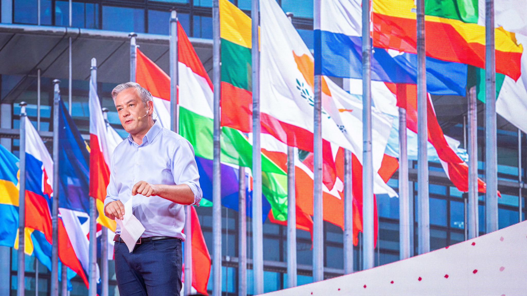 Mężczyzna w jasnoniebieskiej koszuli przemawia ze sceny, trzymając kartkę, na tle licznych flag państwowych.