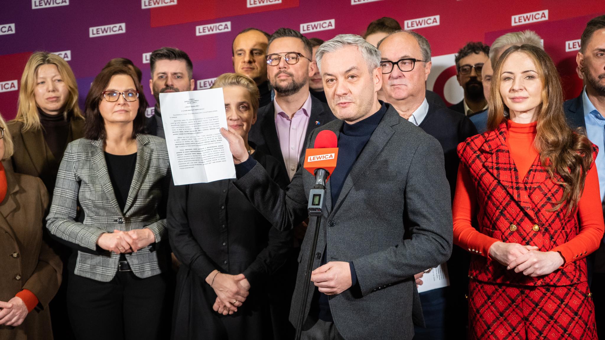 Na zdjęciu przedstawiona jest grupa osób z partii Lewica, z liderem trzymającym dokument i mówiącym do kamery.