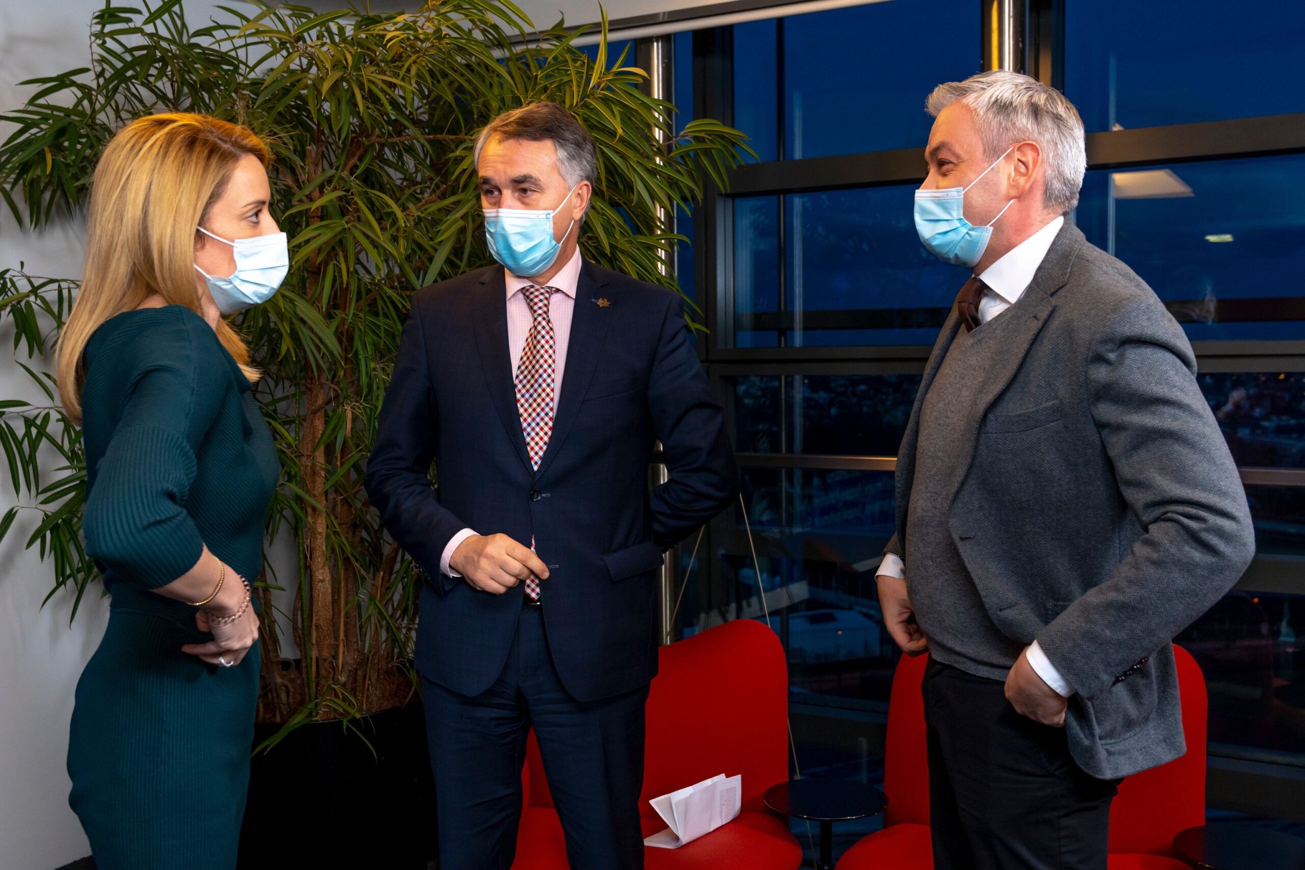 Na zdjęciu trzech ludzi, w tym dwie osoby w maseczkach, rozmawia ze sobą w biurowym wnętrzu z widokiem na miasto w tle.