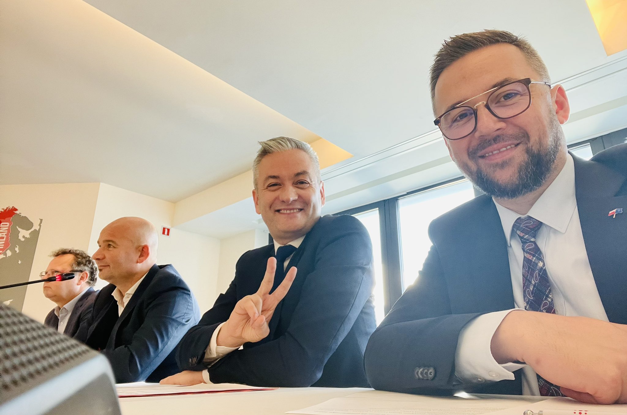 Na zdjęciu widać grupę mężczyzn siedzących przy stole, uśmiechających się i pozujących do zdjęcia, z widocznymi mikrofonami oraz oknem w tle.