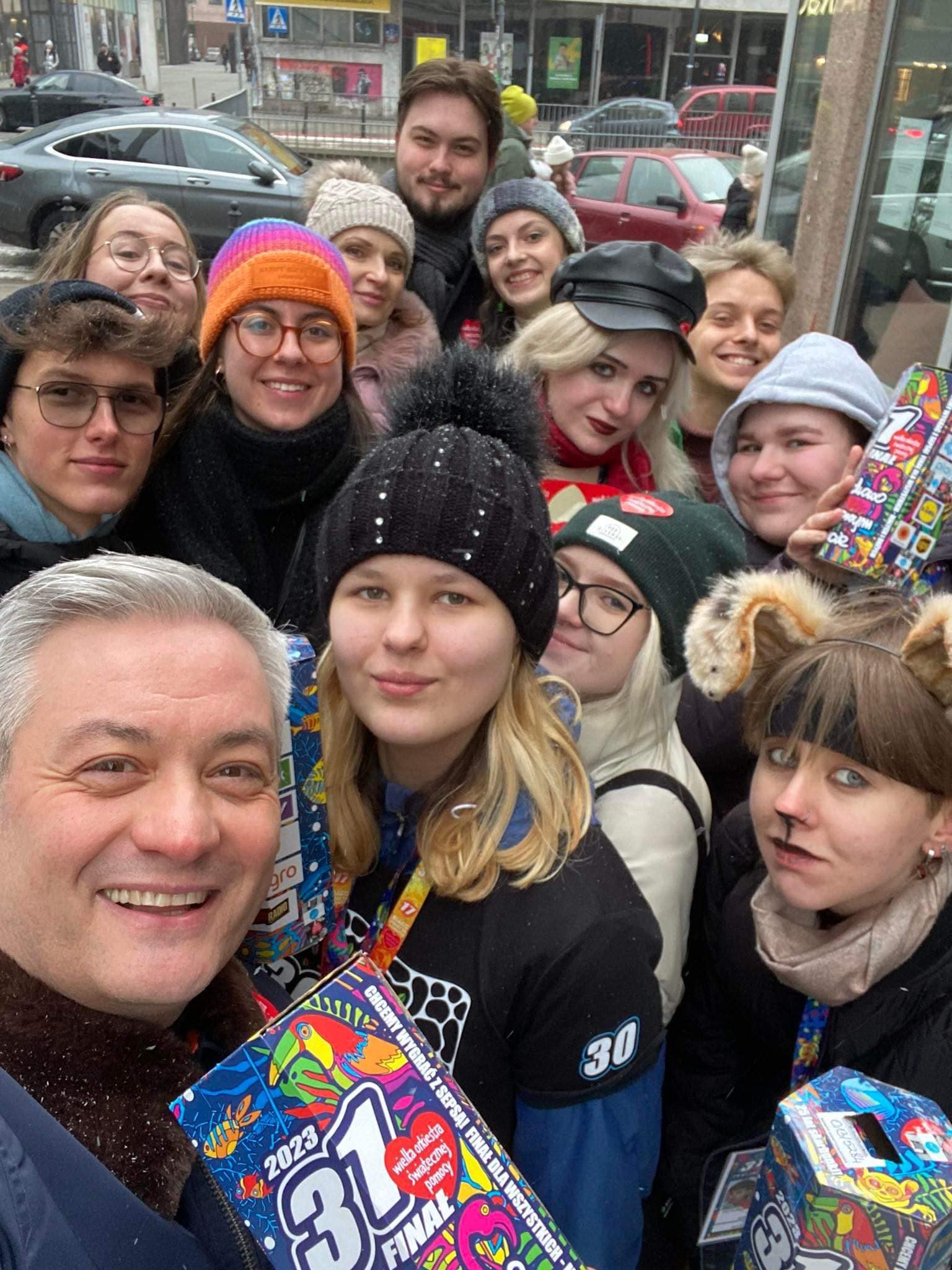 Zdjęcie przedstawia grupę uśmiechniętych ludzi pozującą do selfie, trzymających paczki z kolorowymi grafikami, w zimowej scenerii.