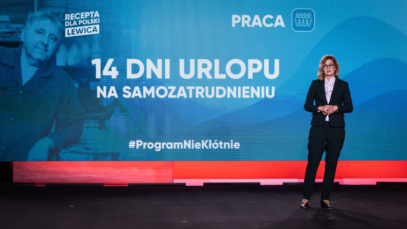 Kobieta w garniturze stoi na scenie przed dużym, niebieskim ekranem z napisem „14 Dni Urlopu na Samozatrudnieniu” oraz logotypami kampanii.