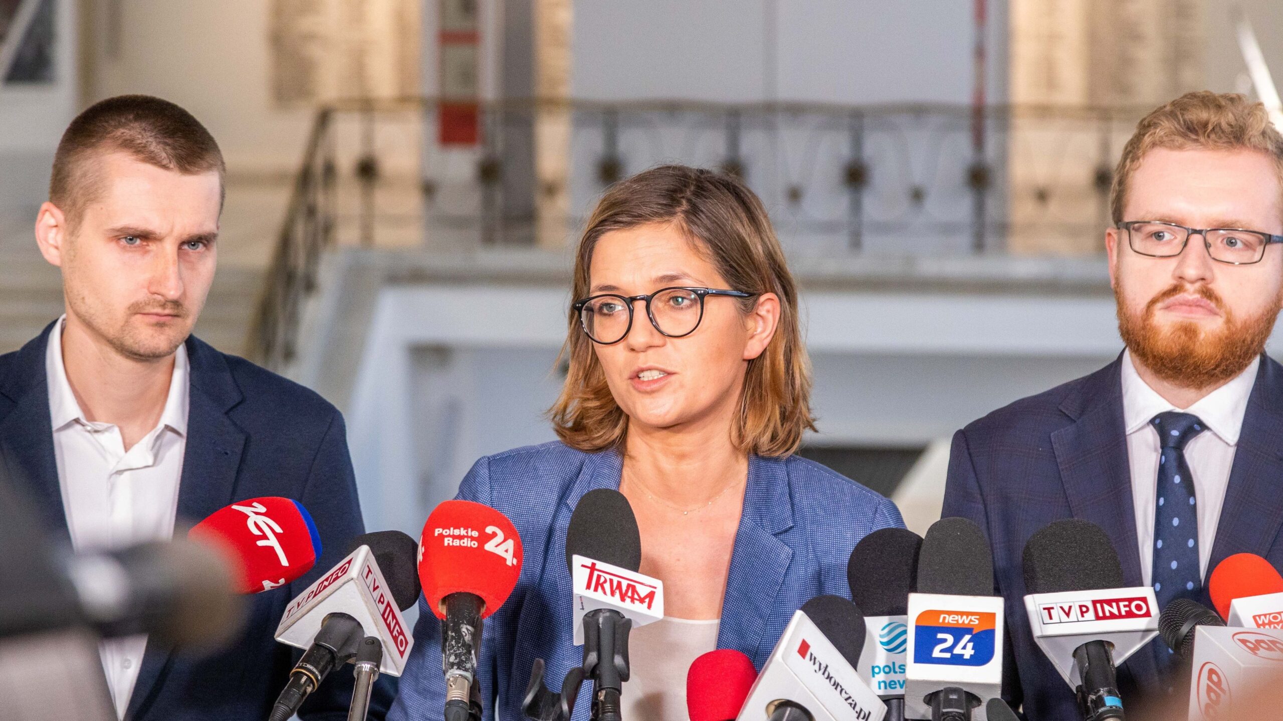 Na zdjęciu widać troje polityków stojących przed mikrofonami podczas konferencji prasowej.