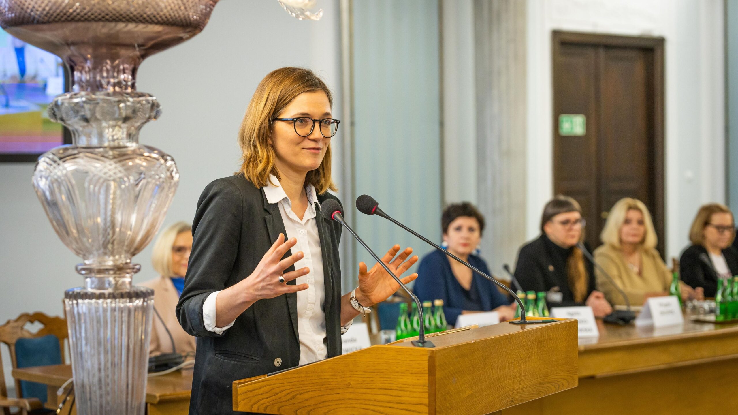 Osoba przemawiająca podczas konferencji, w tle siedzi grupa kobiet, a na stole znajdują się butelki wody.