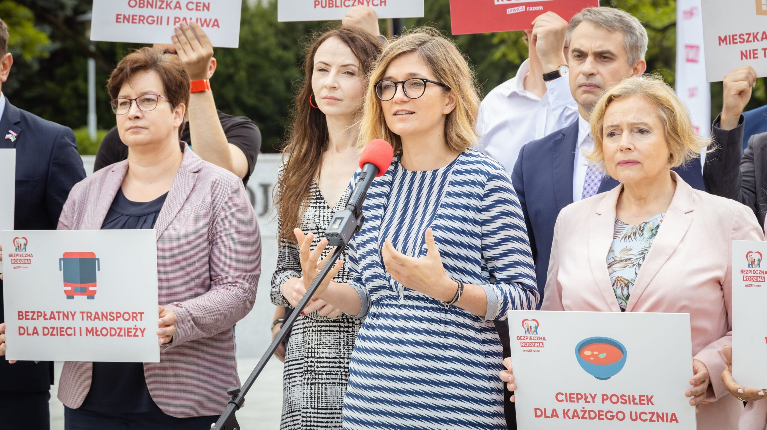 Na zdjęciu widoczni są politycy podczas konferencji prasowej, trzymający transparenty dotyczące ważnych kwestii społecznych, takich jak transport dla dzieci i ceny energii.