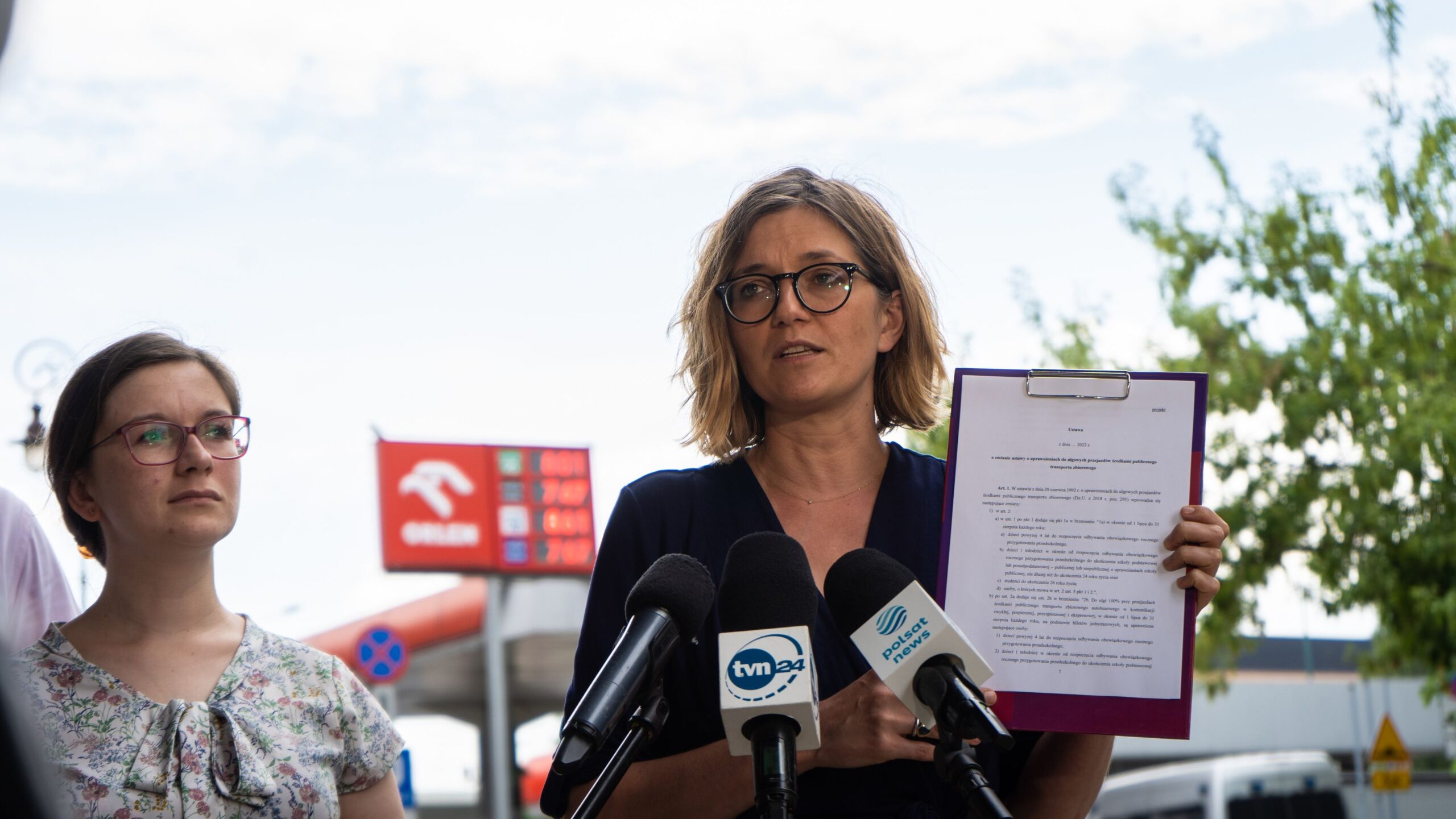 Na zdjęciu widać kobietę z krótkimi włosami, noszącą okulary, która przemawia podczas konferencji prasowej, trzymając w ręku dokument, obok niej stoi druga kobieta.