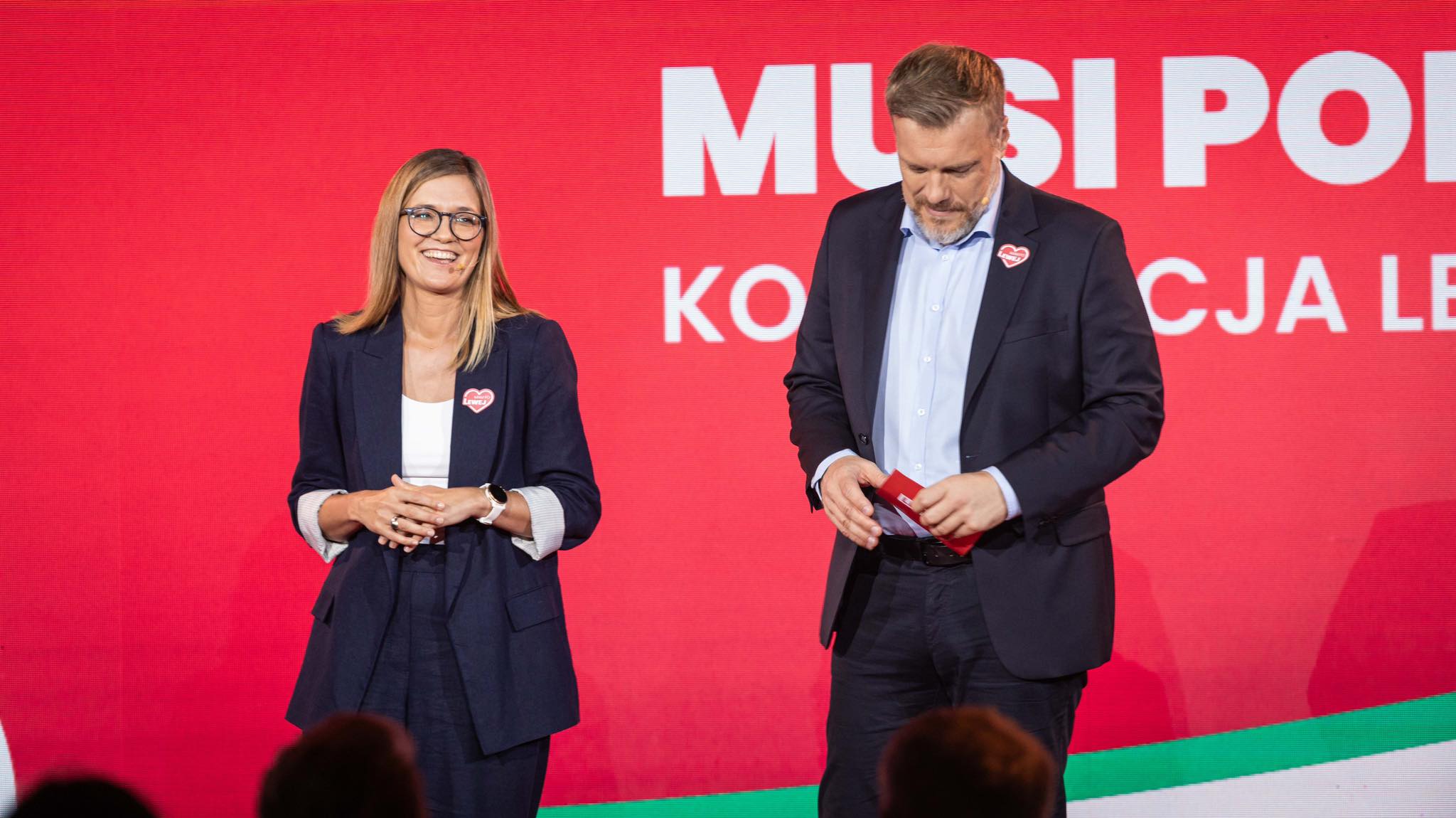 Dwoje osób stojących na scenie w garniturach: kobieta po lewej uśmiecha się, mężczyzna po prawej patrzy w dół, a w tle w czerwonym ekranie widnieją białe napisy „Uczciwa praca musi popłacać”.