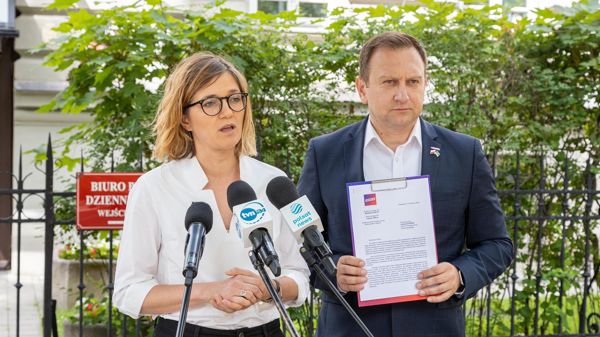 Na zdjęciu widać dwoje mówiących ludzi przy mikrofonach, trzymających dokumenty, w tle rosnące drzewa i znak