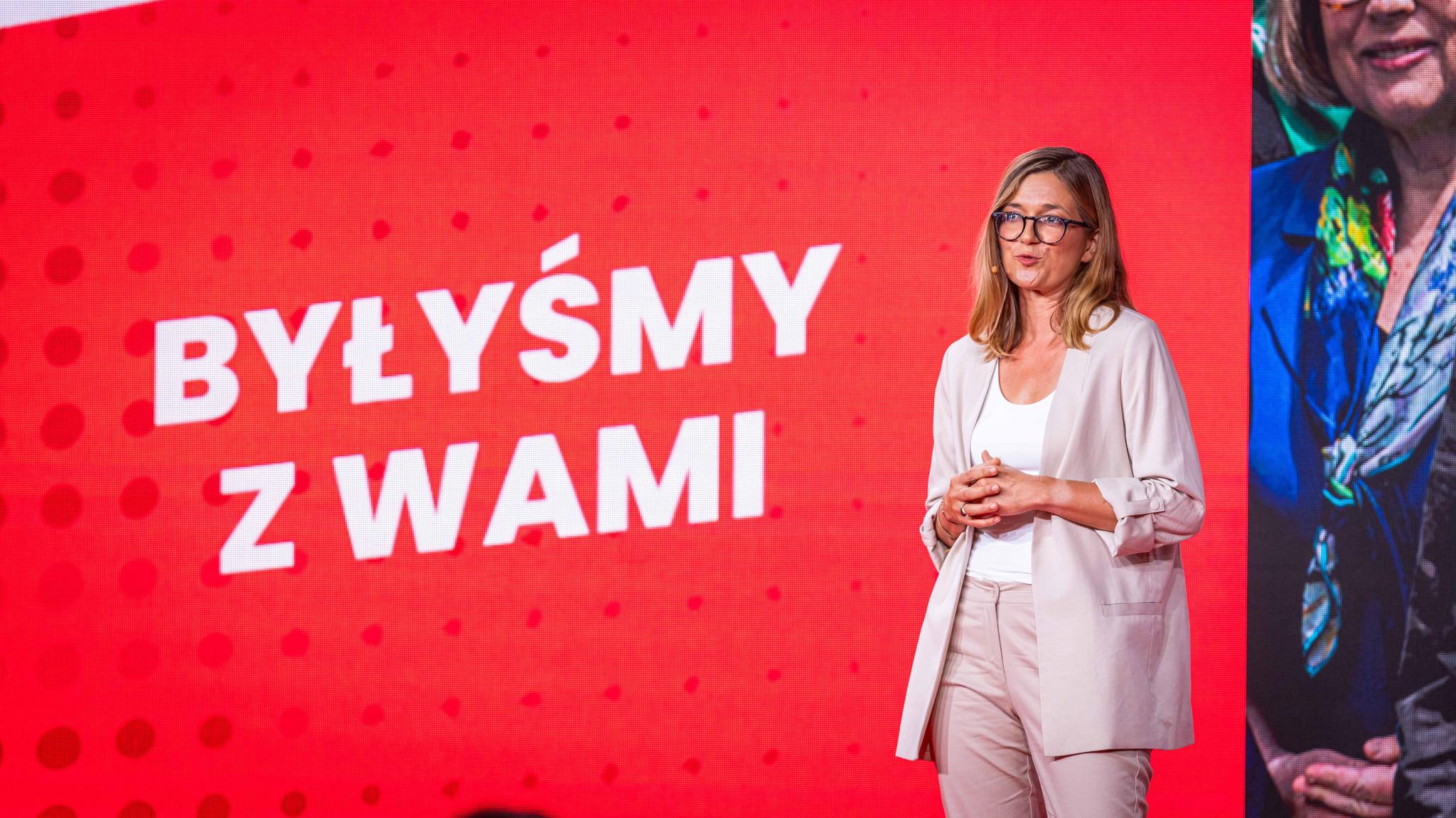 Kobieta w jasnym garniturze i okularach stoi na scenie przed czerwonym tłem z napisem „BYŁYŚMY Z WAMI”.