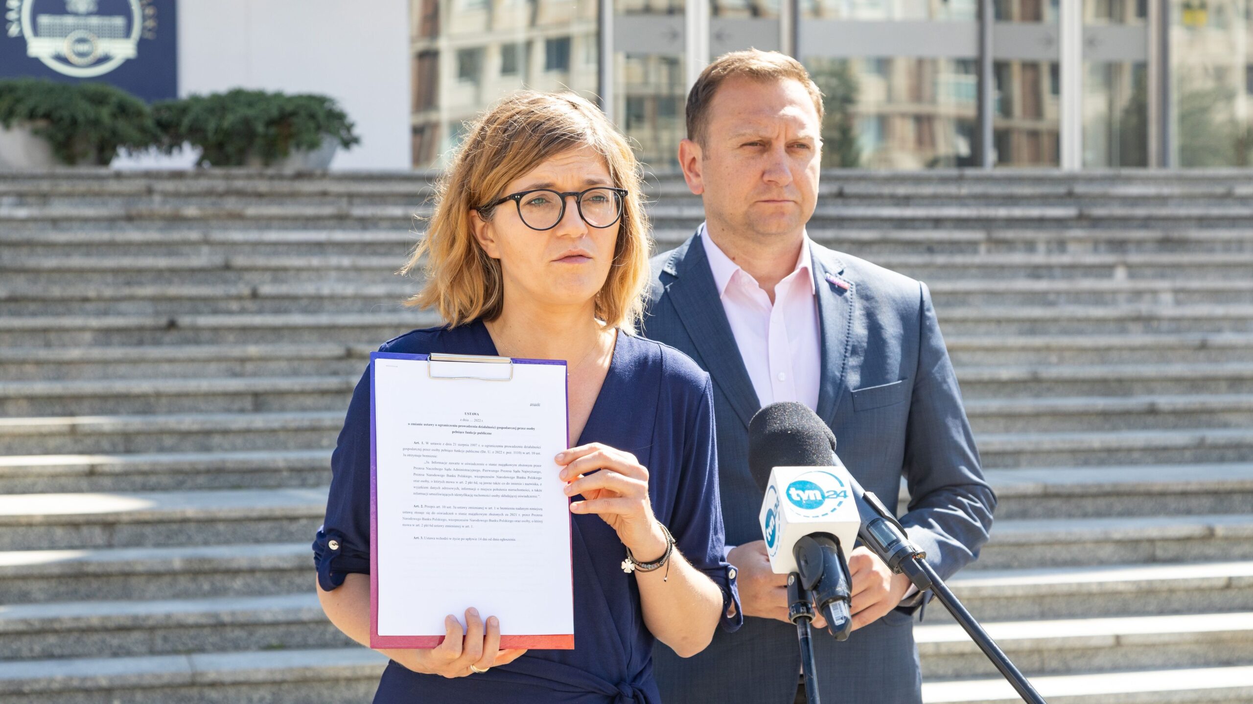 Na zdjęciu widoczna jest kobieta trzymająca dokument oraz mężczyzna w garniturze, oboje stoją przed budynkiem podczas konferencji prasowej.