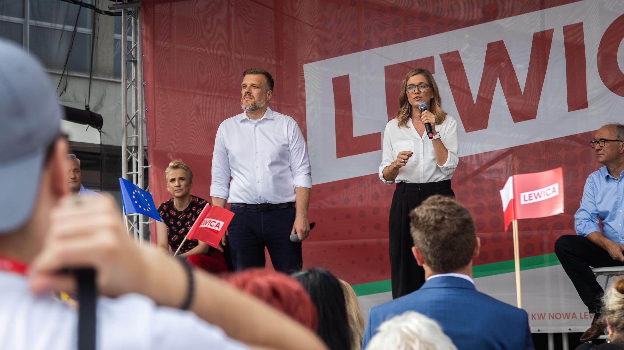 Kobieta z okularami przemawia do tłumu na wiecu Lewicy w Częstochowie, stojąc obok mężczyzny; w tle widoczny czerwony baner z napisem Lewica i flagi.