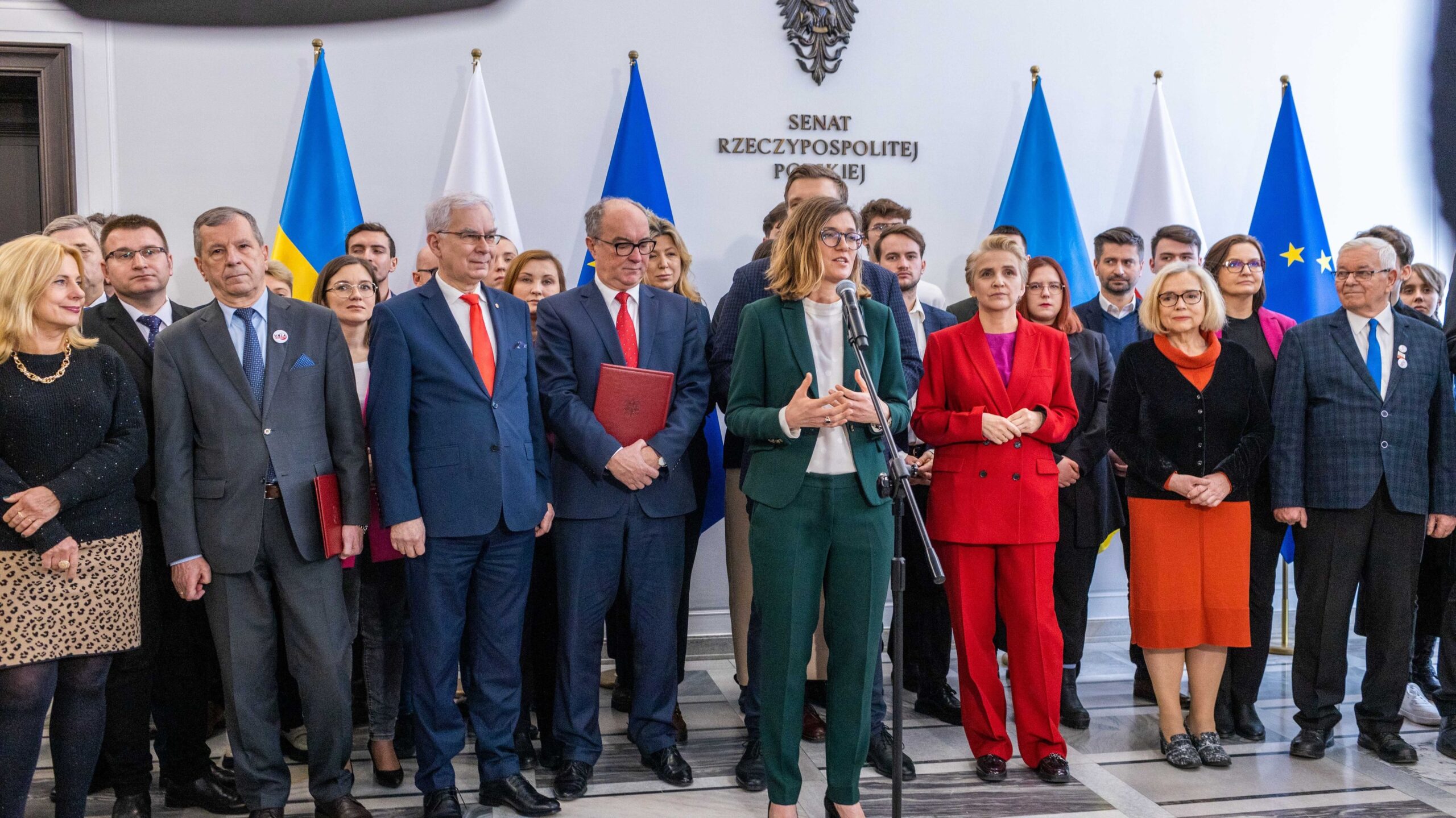 Grupa polityków lewicy podczas podpisywania porozumienia; kobieta w zielonym garniturze przemawia do mikrofonu, a za nią stoi reszta uczestników, w tle widoczne flagi Polski, Ukrainy i Unii Europejskiej.