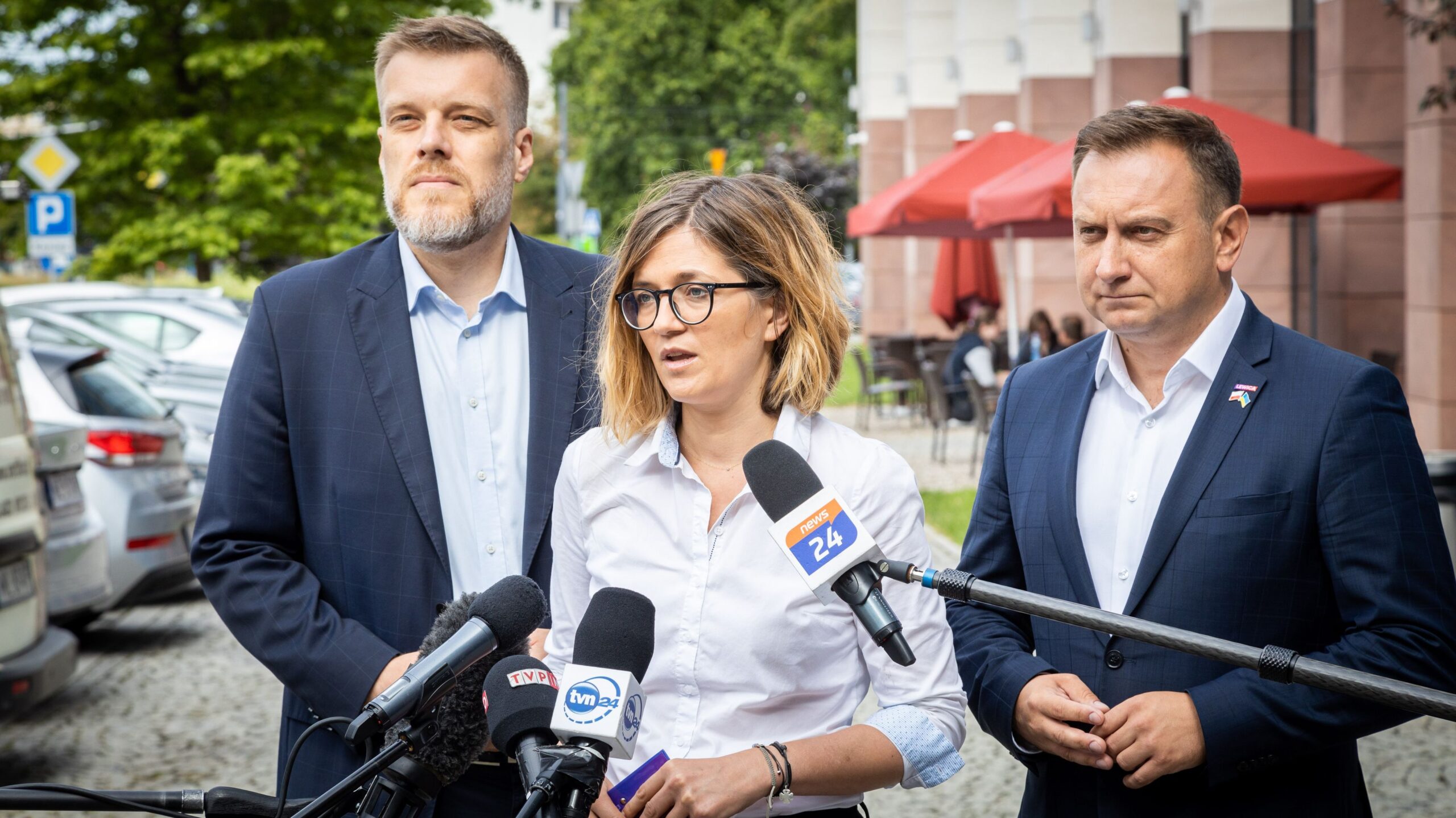 Na zdjęciu znajdują się trzy osoby podczas konferencji prasowej, z mikrofonami i kamerami mediów w tle.