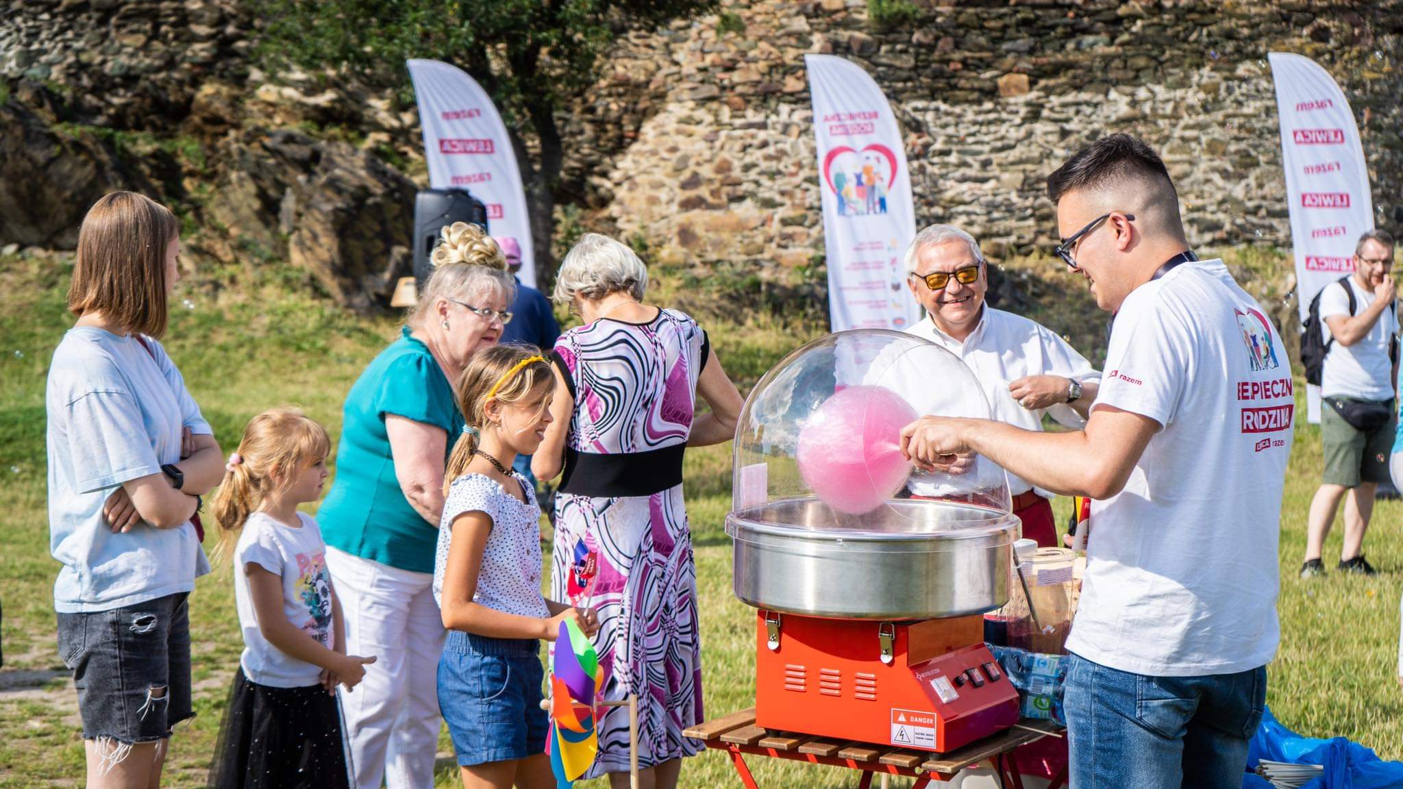 Na zdjęciu widać grupę ludzi zgromadzonych wokół maszyny do waty cukrowej, w której chłopcy i dziewczynki cieszą się słodkim przysmakiem podczas plenerowej imprezy.