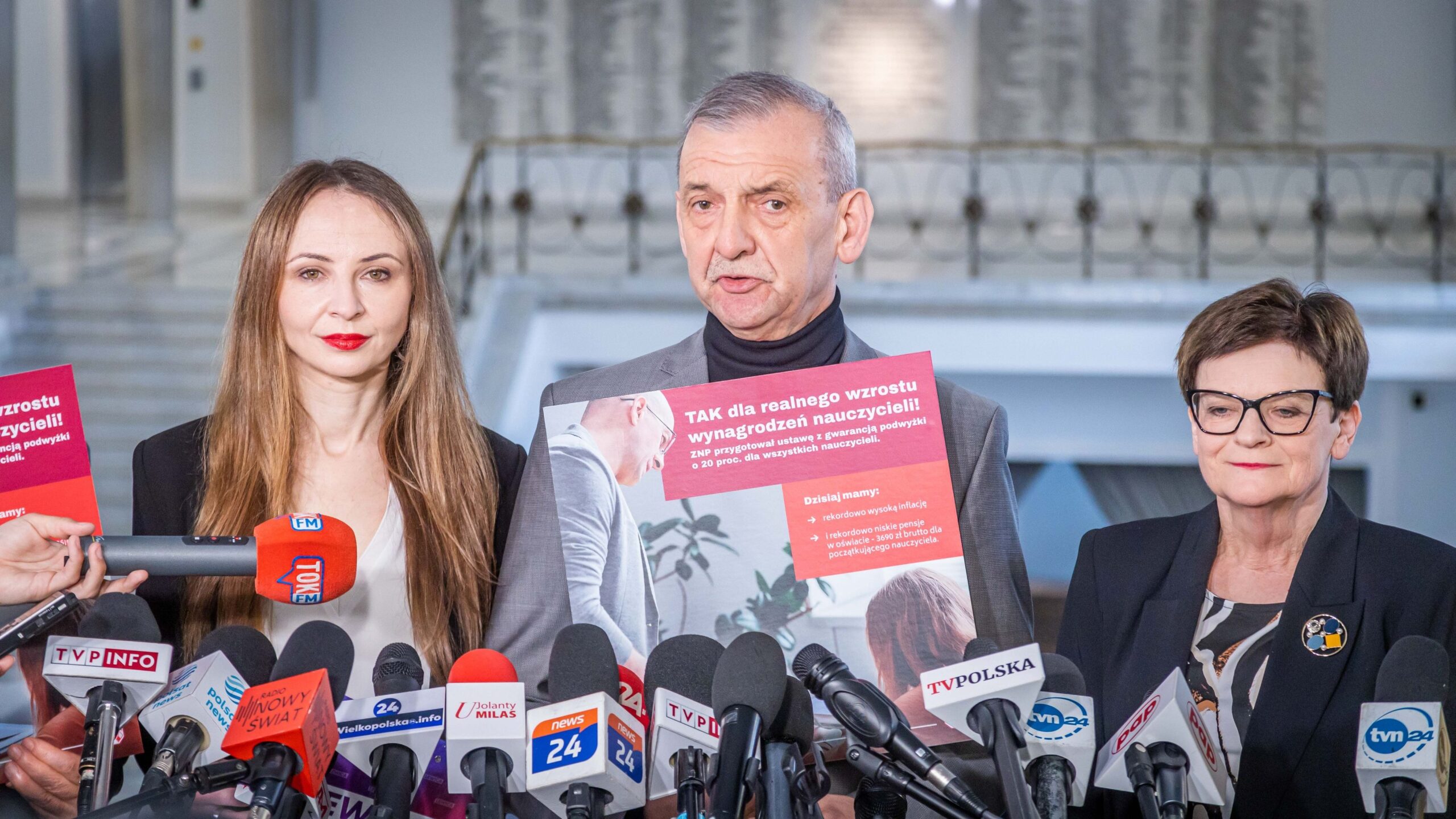Trzy osoby stojące na konferencji prasowej z licznymi mikrofonami; w centrum mężczyzna trzyma plakat dotyczący 20-procentowej podwyżki dla nauczycieli w ramach inicjatywy ZNP i demokratycznej opozycji.