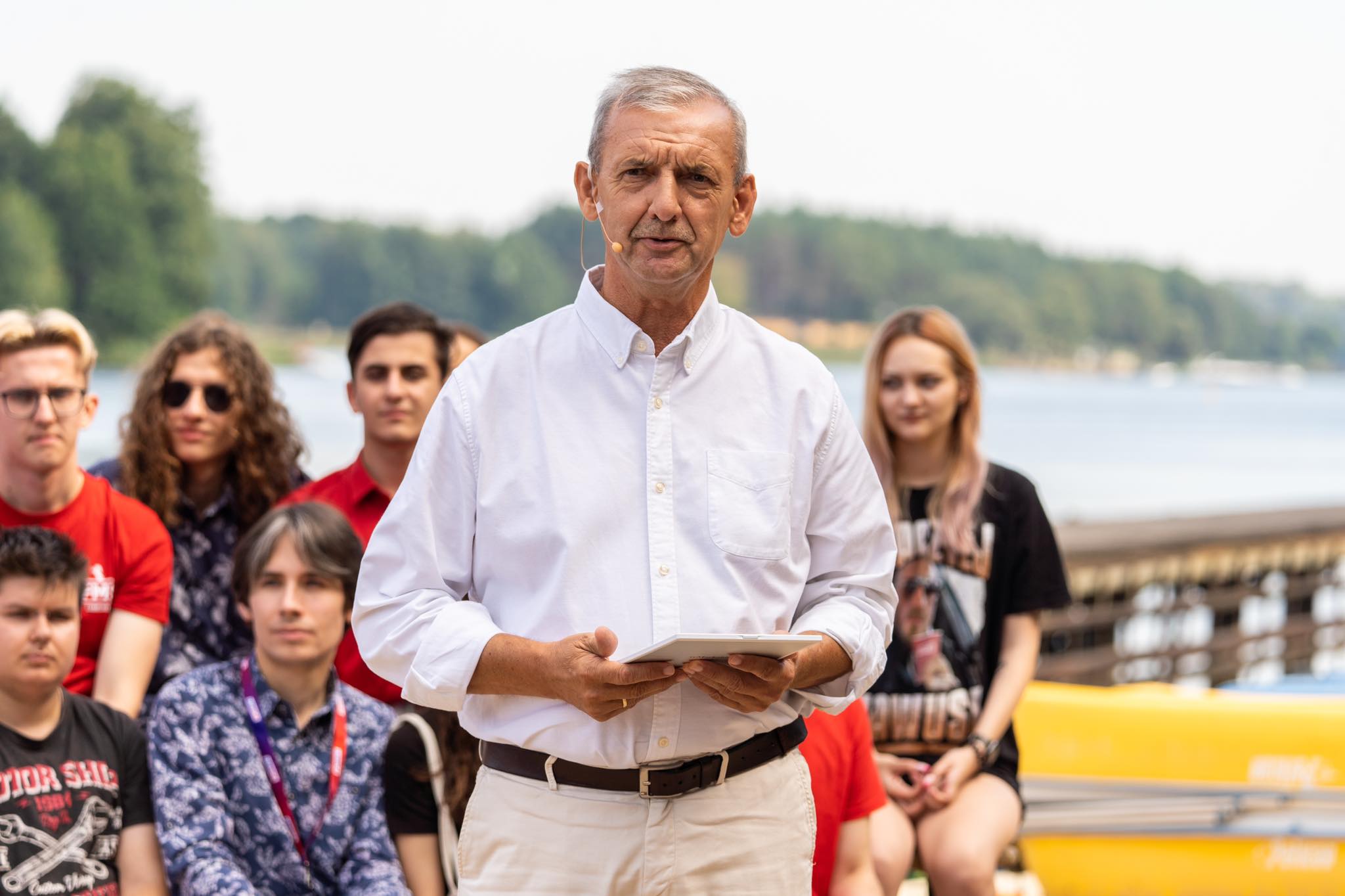Mężczyzna w białej koszuli z mikrofonem na uchu przemawia podczas konwencji programowej Lewicy, stojąc przed grupą młodych osób nad jeziorem.