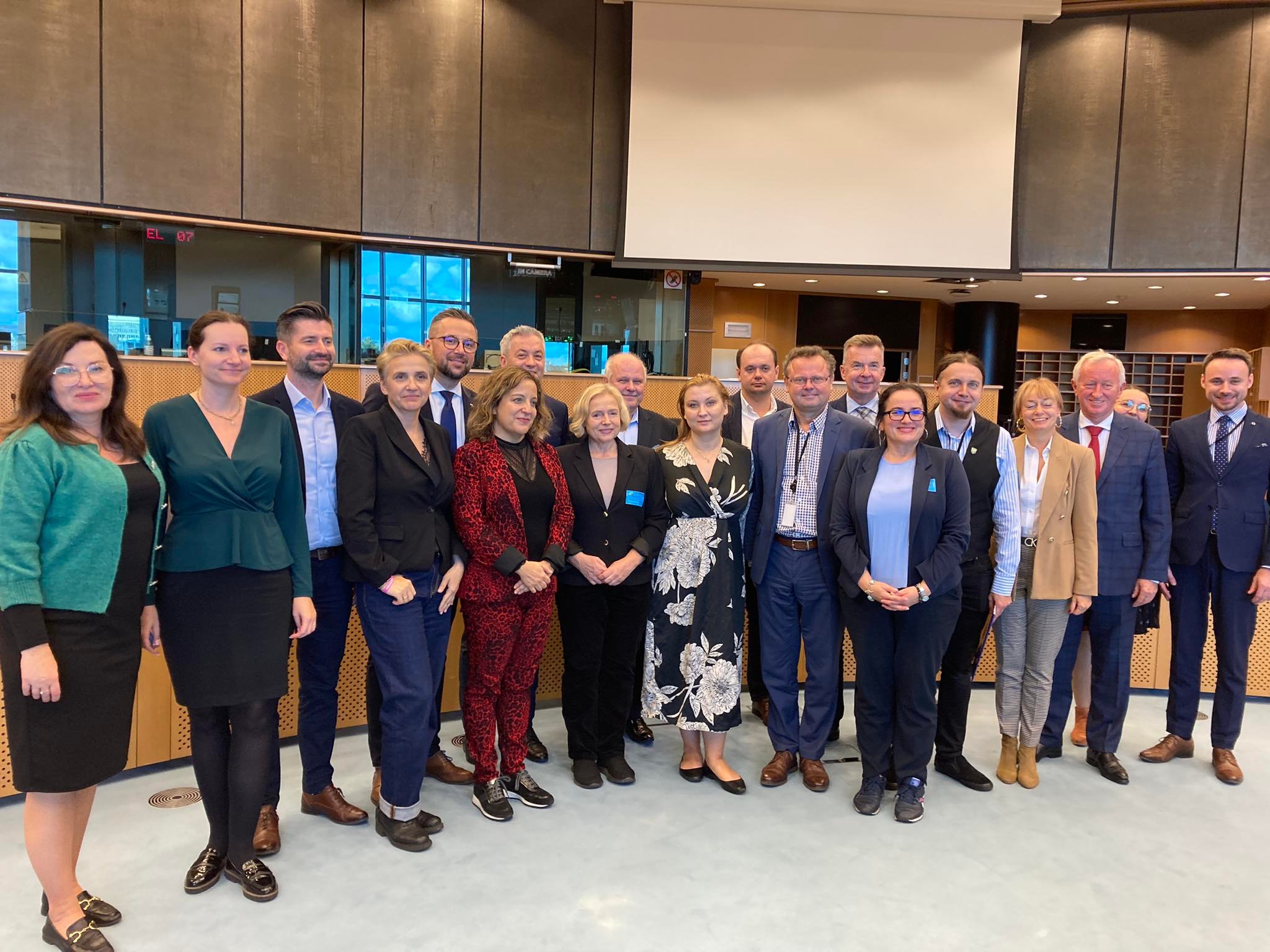 Grupa ludzi w formalnym stroju stoi razem do zdjęcia w sali konferencyjnej z dużymi oknami.