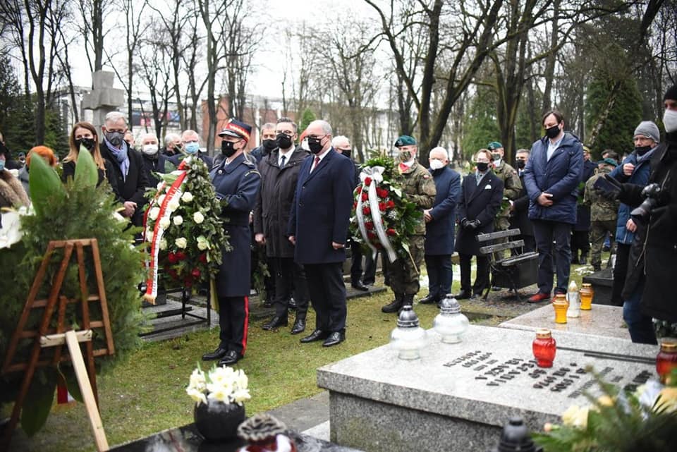 Grupa osób, w mundurach i cywilach, składa wieńce i zapala znicze na cmentarzu podczas uroczystości pożegnalnej.