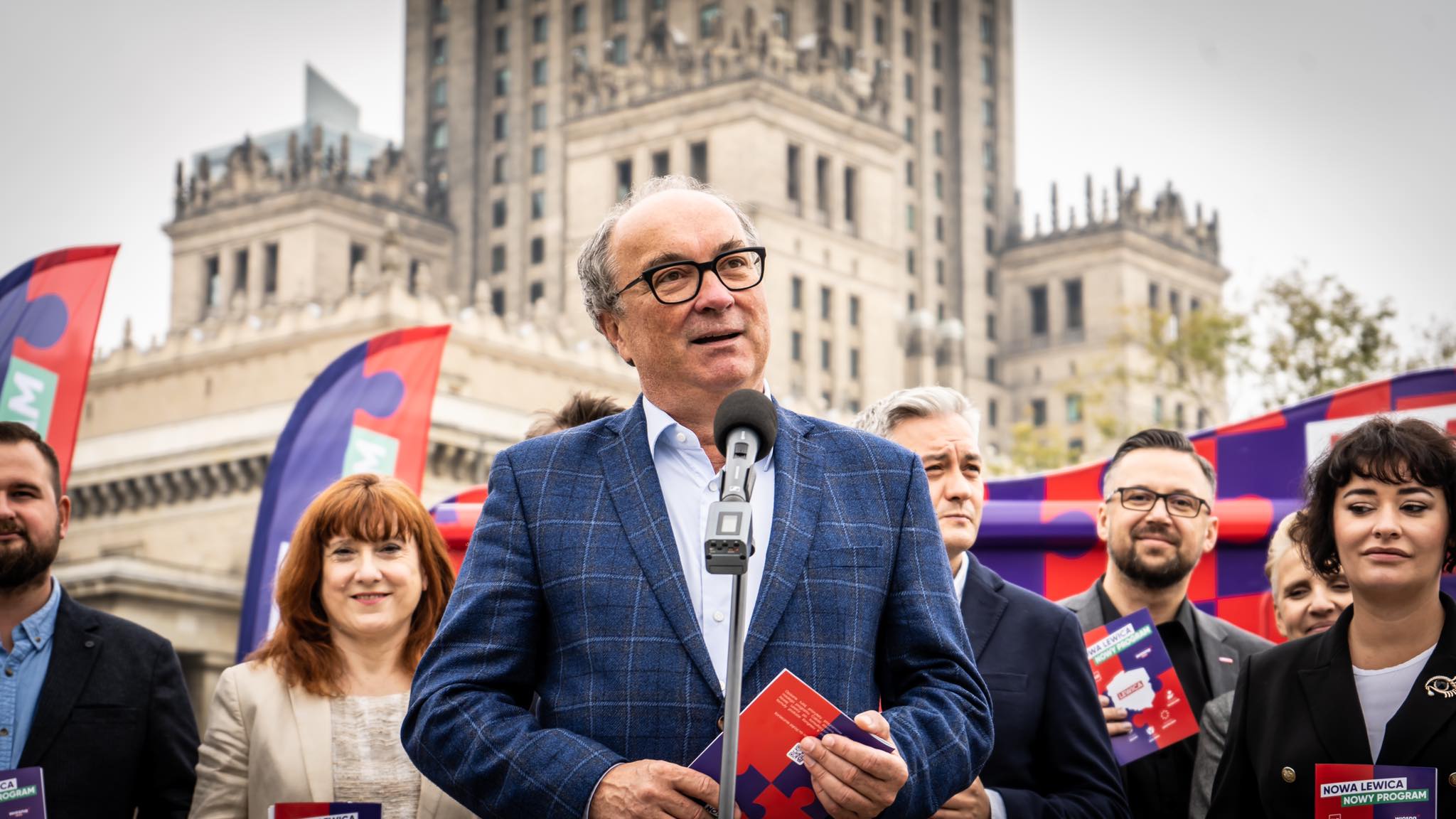 Mężczyzna w granatowo-wzorzystym garniturze przemawia do mikrofonu podczas konferencji Nowej Lewicy, otoczony przez współpracowników na tle zabytkowej architektury.