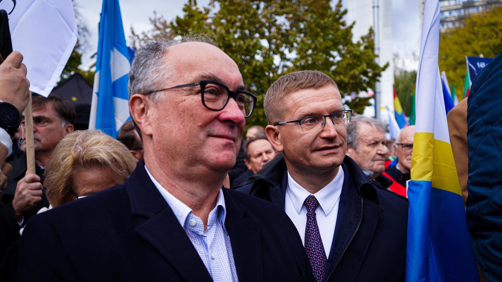 Na zdjęciu przedstawieni są mężczyźni w okularach, którzy uczestniczą w wydarzeniu publicznym, otoczeni przez innych ludzi i flagi.