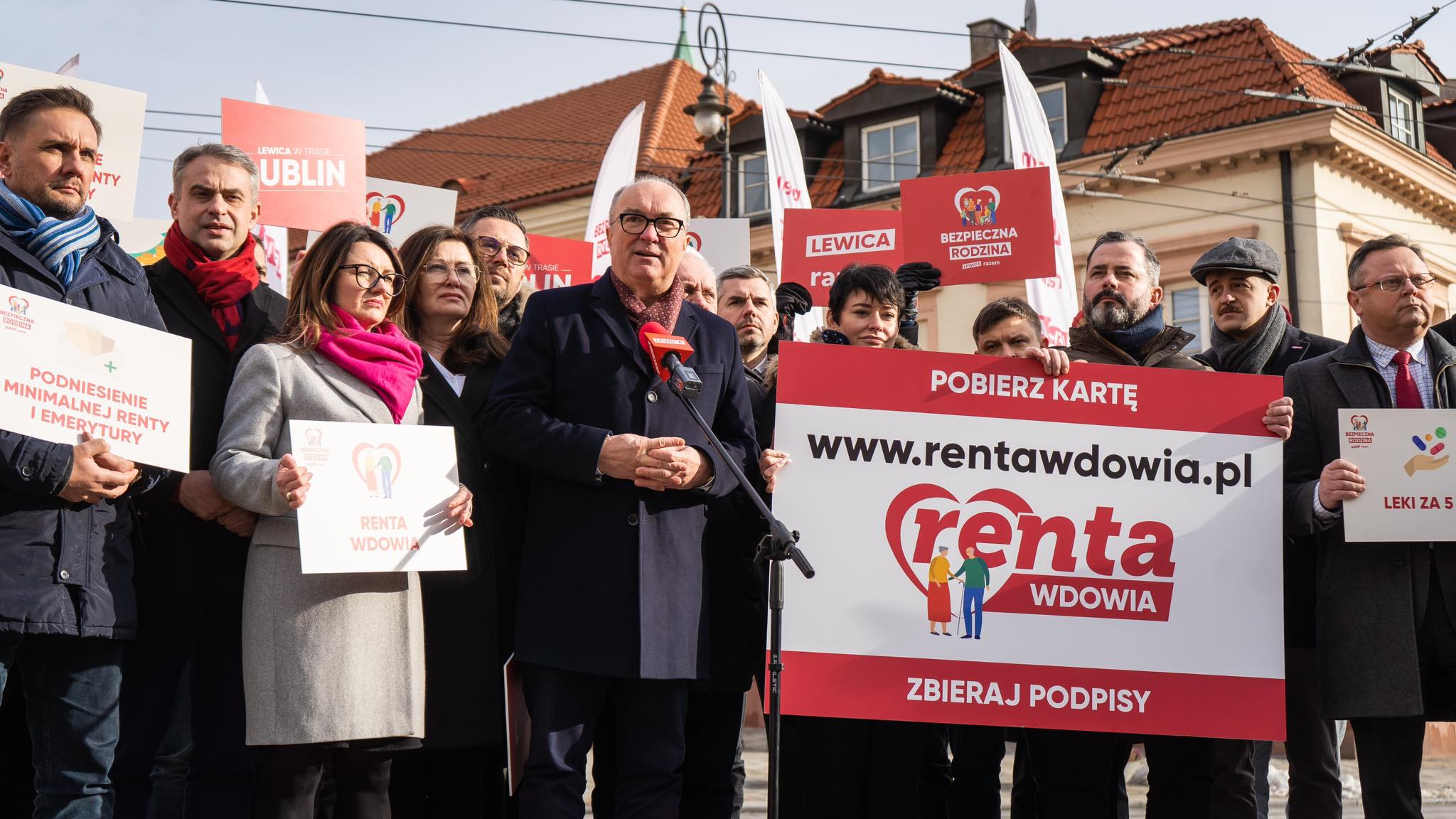 Na zdjęciu przedstawiono grupę osób, które uczestniczą w wydarzeniu promującym zbieranie podpisów pod inicjatywą dotyczącą renty wdowiej, trzymając transparenty i banery.