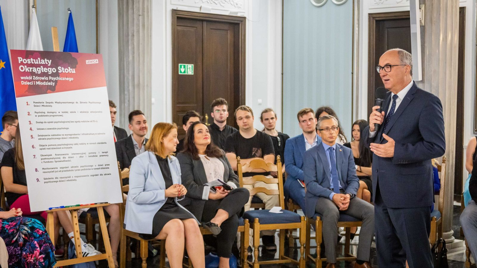 Mężczyzna w garniturze przemawia do publiczności podczas konferencji o zdrowiu psychicznym dzieci i młodzieży; w tle stoi plansza z postulatami Okrągłego Stołu.