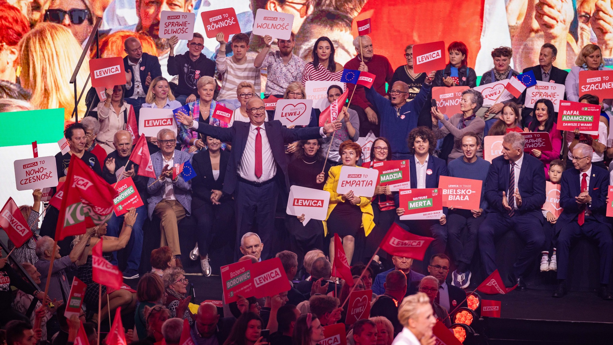 Grupa uczestników konwencji Lewicy w Będzinie na scenie trzyma plakaty i czerwone flagi, a w centrum znajduje się mężczyzna z szeroko rozłożonymi ramionami.