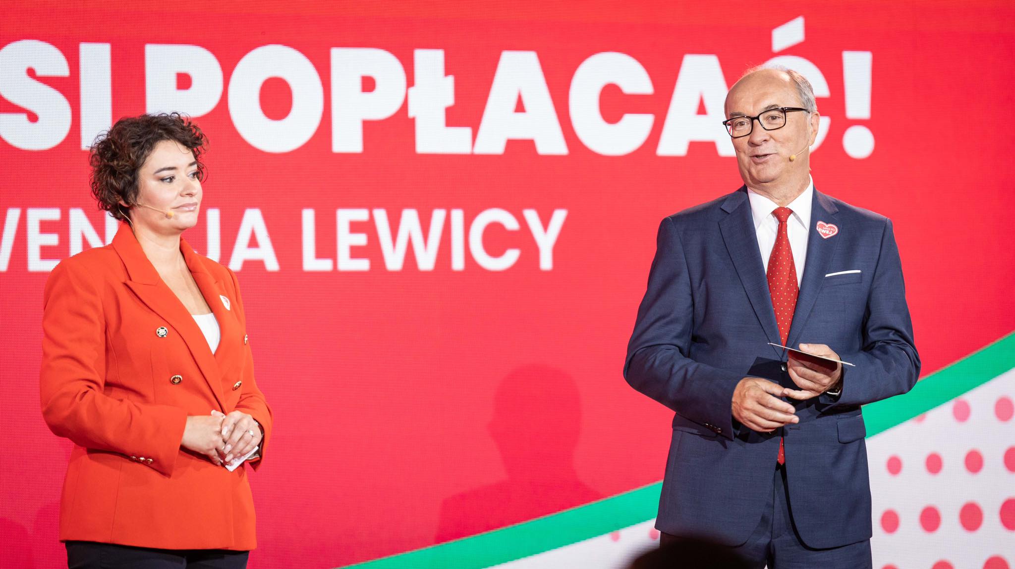 Dwie osoby stoją na scenie podczas wydarzenia; kobieta w czerwonej marynarce i mężczyzna w granatowym garniturze, na tle dużego czerwonego banera z białymi napisami.