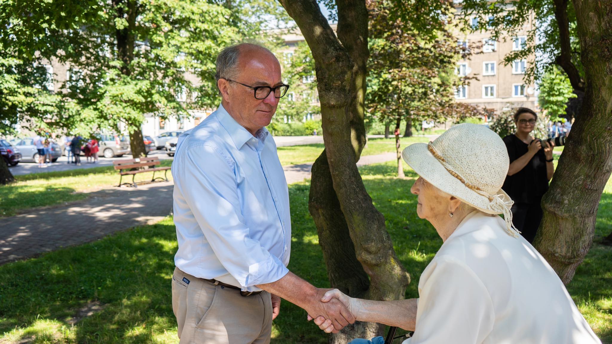 Mężczyzna w jasnej koszuli i okularach ściska dłoń starszej kobiecie w kapeluszu, stojąc w parku; w tle widać inne osoby i osobę z aparatem.