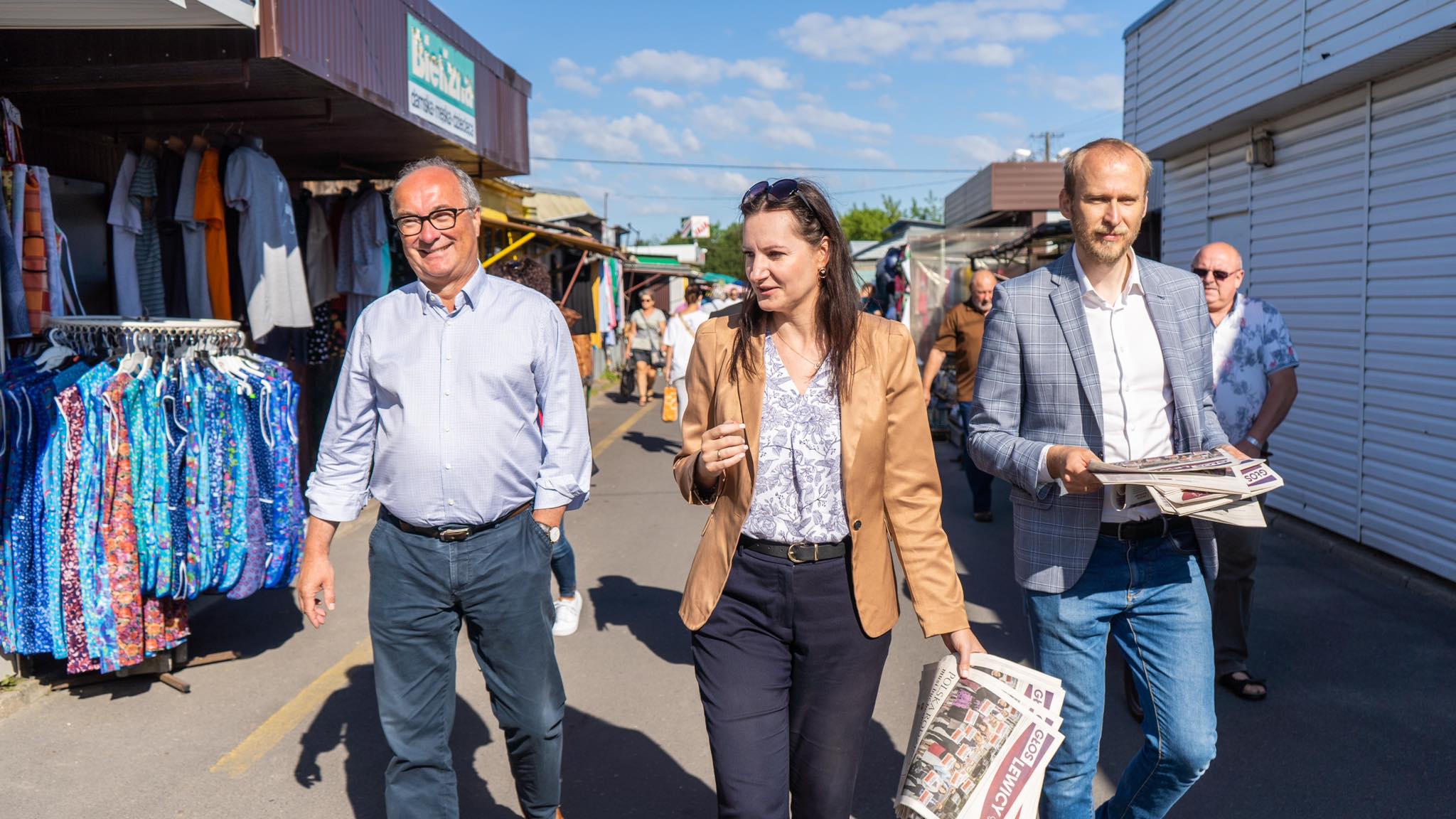 Trójka dorosłych spaceruje po targu z kolorowymi ubraniami na wieszakach, kobieta w beżowym żakiecie rozgląda się, a mężczyzna w kratkowanej marynarce trzyma gazety.