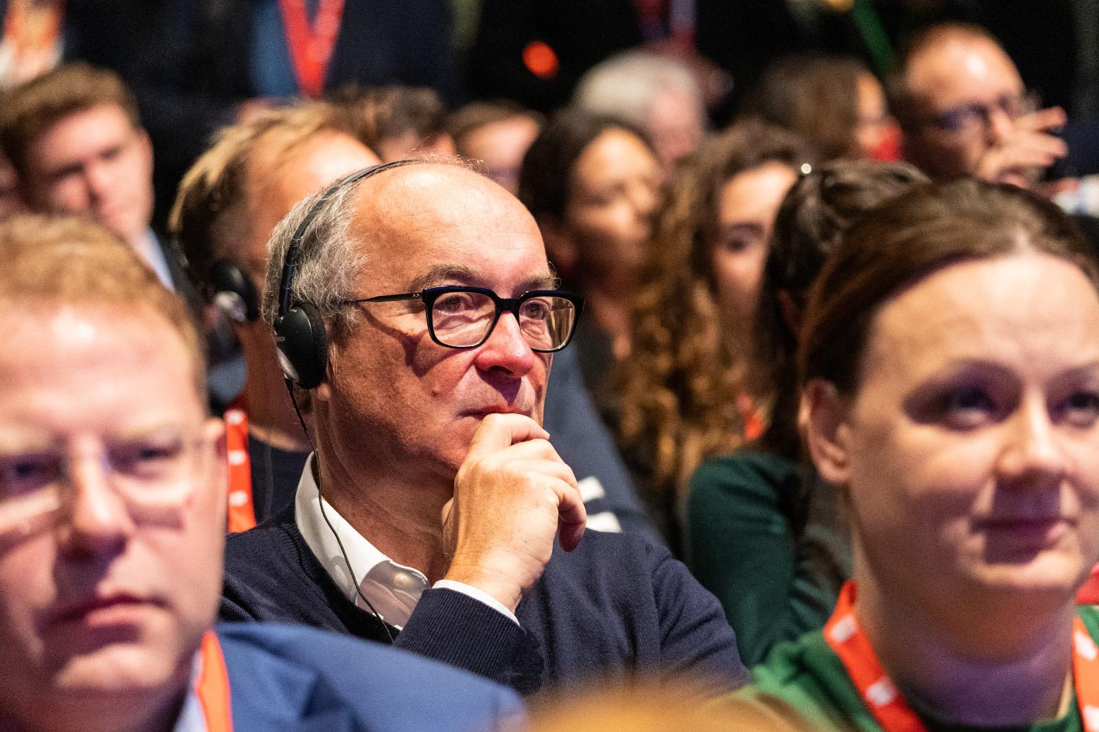 Na zdjęciu widać grupę osób, z których jedna w słuchawkach intensywnie słucha, zastanawiając się nad prezentacją lub dyskusją.