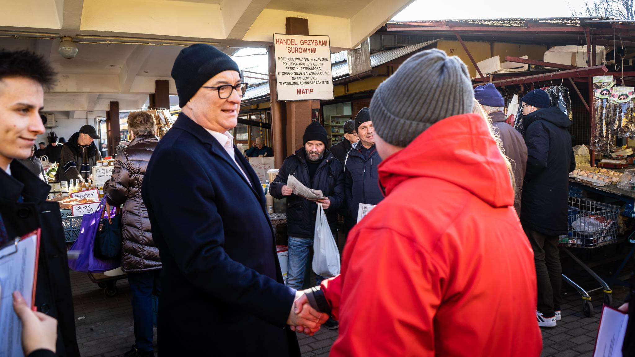 Na zdjęciu widoczna jest scena targowa, gdzie mężczyzna w okularach i czapce uścisnął dłoń osobie w czerwonym płaszczu, a w tle widać innych ludzi oraz stoiska z artykułami spożywczymi.