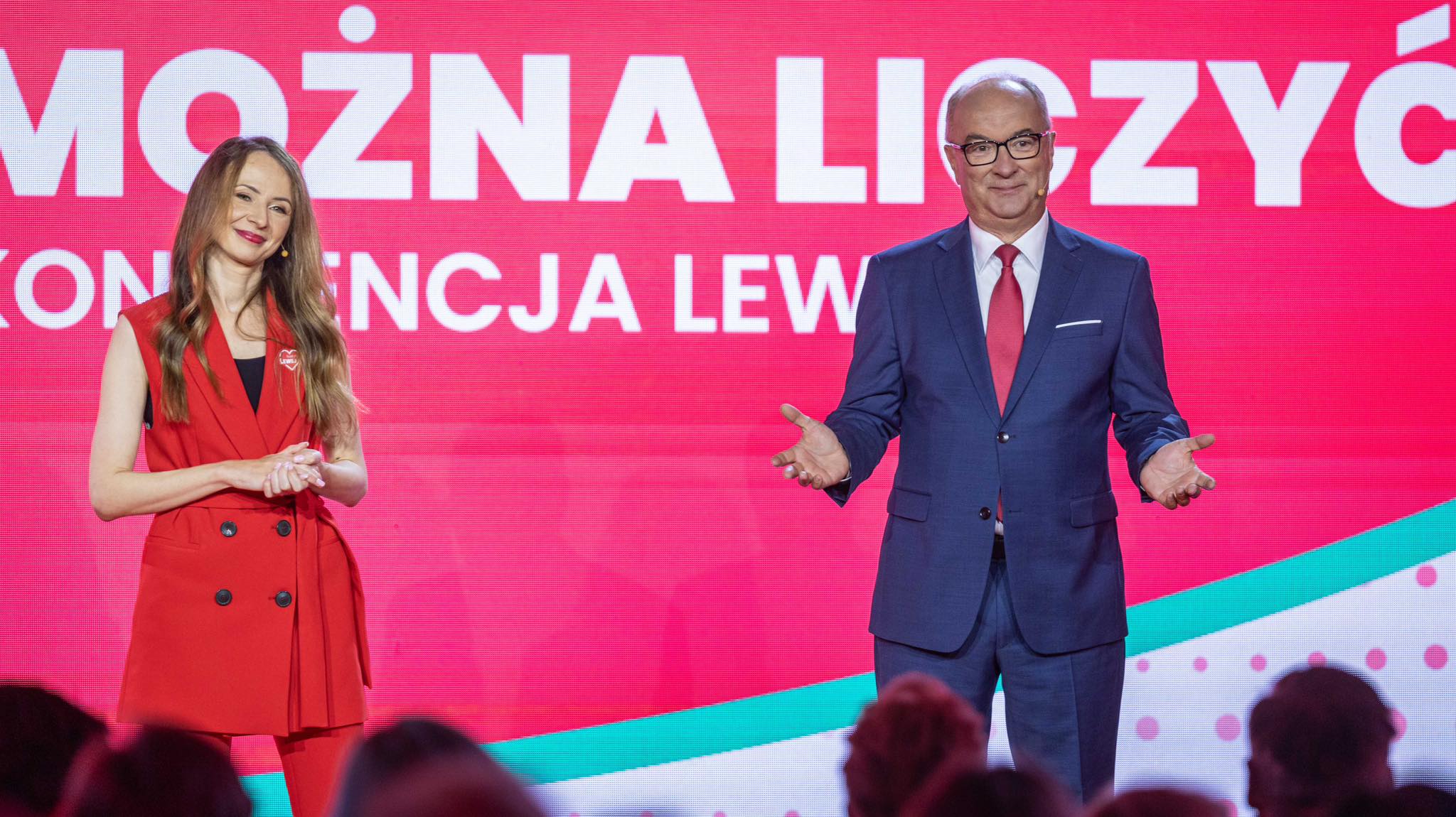 Na scenie stoją kobieta w czerwonej sukience i mężczyzna w granatowym garniturze z czerwonym krawatem, przed dużym różowym ekranem z napisem „MOŻNA LICZYĆ”.