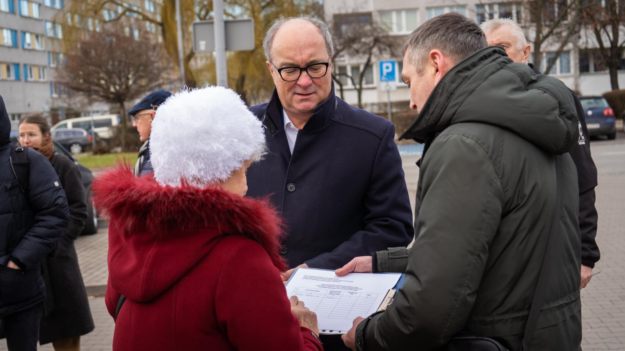 Grupa dorosłych rozmawia na chodniku w mieście; kobieta w czerwonej kurtce z białym, futrzanym kapeluszem trzyma dokumenty.