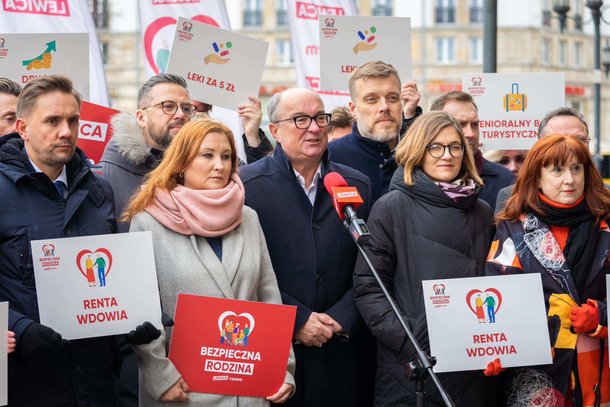 Na zdjęciu grupa osób trzyma transparenty z hasłami dotyczącymi polityki społecznej, podczas konferencji prasowej na zewnątrz.