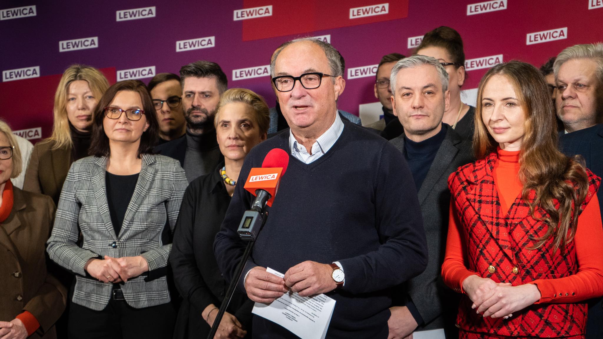 Na zdjęciu widoczna jest grupa polityków z partii Lewica, z liderem mówiącym do mikrofonu w środku, na tle fioletowego plakatu.